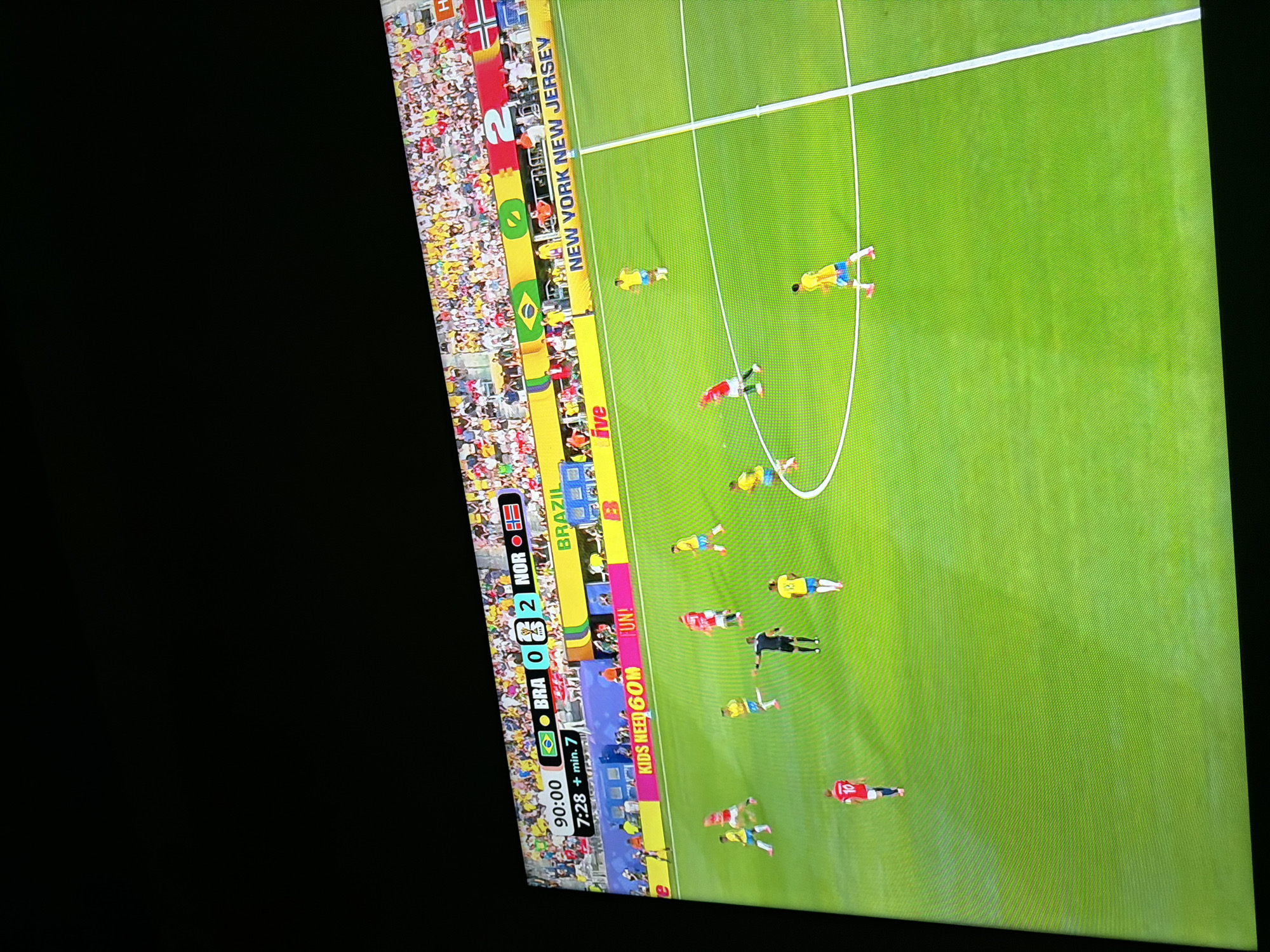 The image shows a screen displaying a soccer match. The score is BRA 0 - 2 NOR. The game clock shows 90:00 and 7:28 + min. 7. The players are wearing yellow and red uniforms. The stadium appears to be full of spectators. The text "BRAZIL" and "NEW YORK NEW JERSEY" are visible on a banner in the background. The scene is of a soccer game broadcast on a screen.