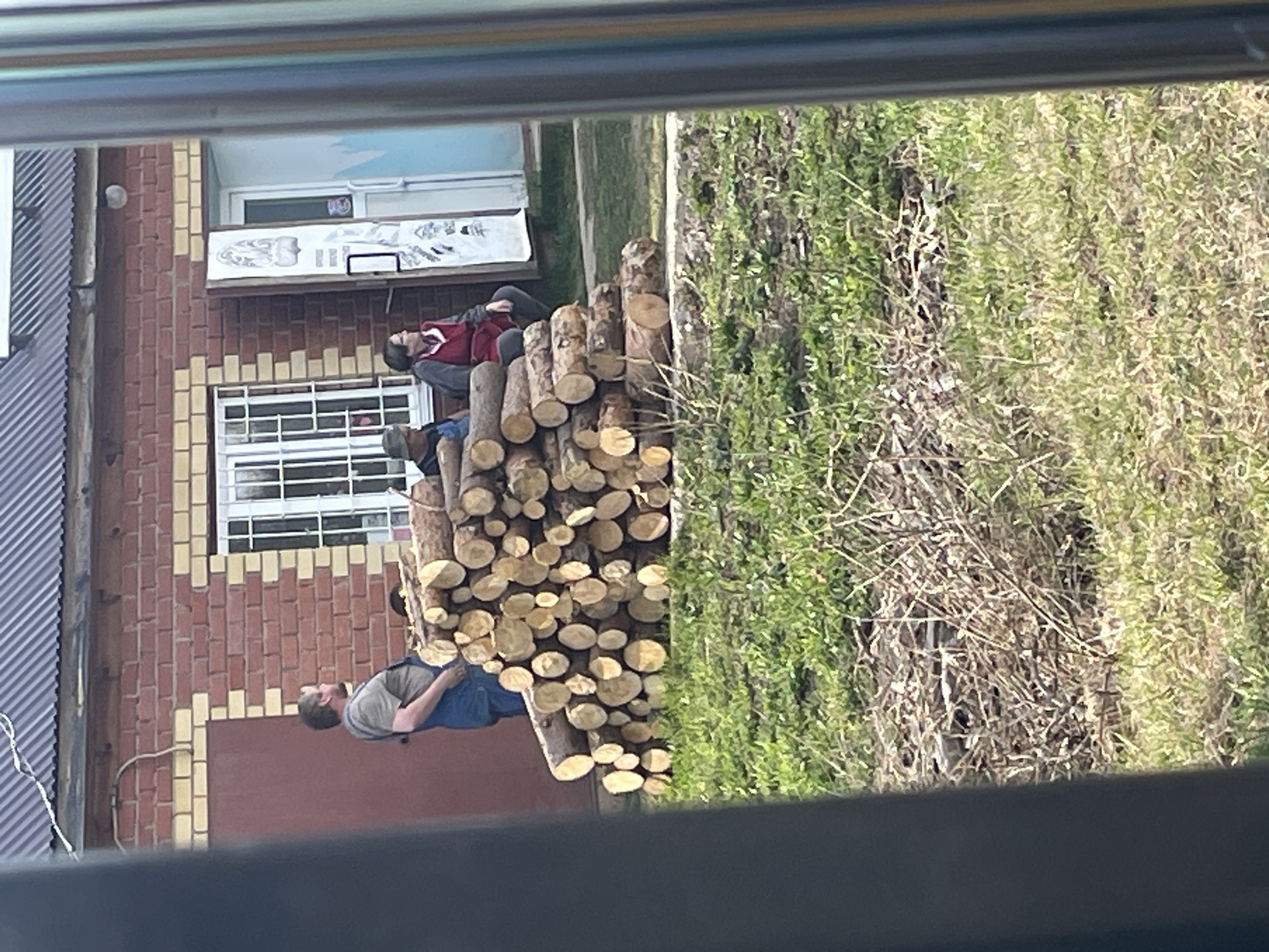 The image, likely taken from inside a building looking out through a window in Поедуги, Russia, shows a stack of firewood logs adjacent to a building's exterior wall of red and yellow brick. Three individuals are positioned atop the log pile. The uppermost person wears a red and gray jacket. The second person is dressed in blue with a dark colored shirt, and the lowermost individual wears blue overalls and a gray shirt. A patch of vegetation is visible behind the logs. A metal roof is seen on the left and top left corners. The scene suggests a rural or suburban setting.