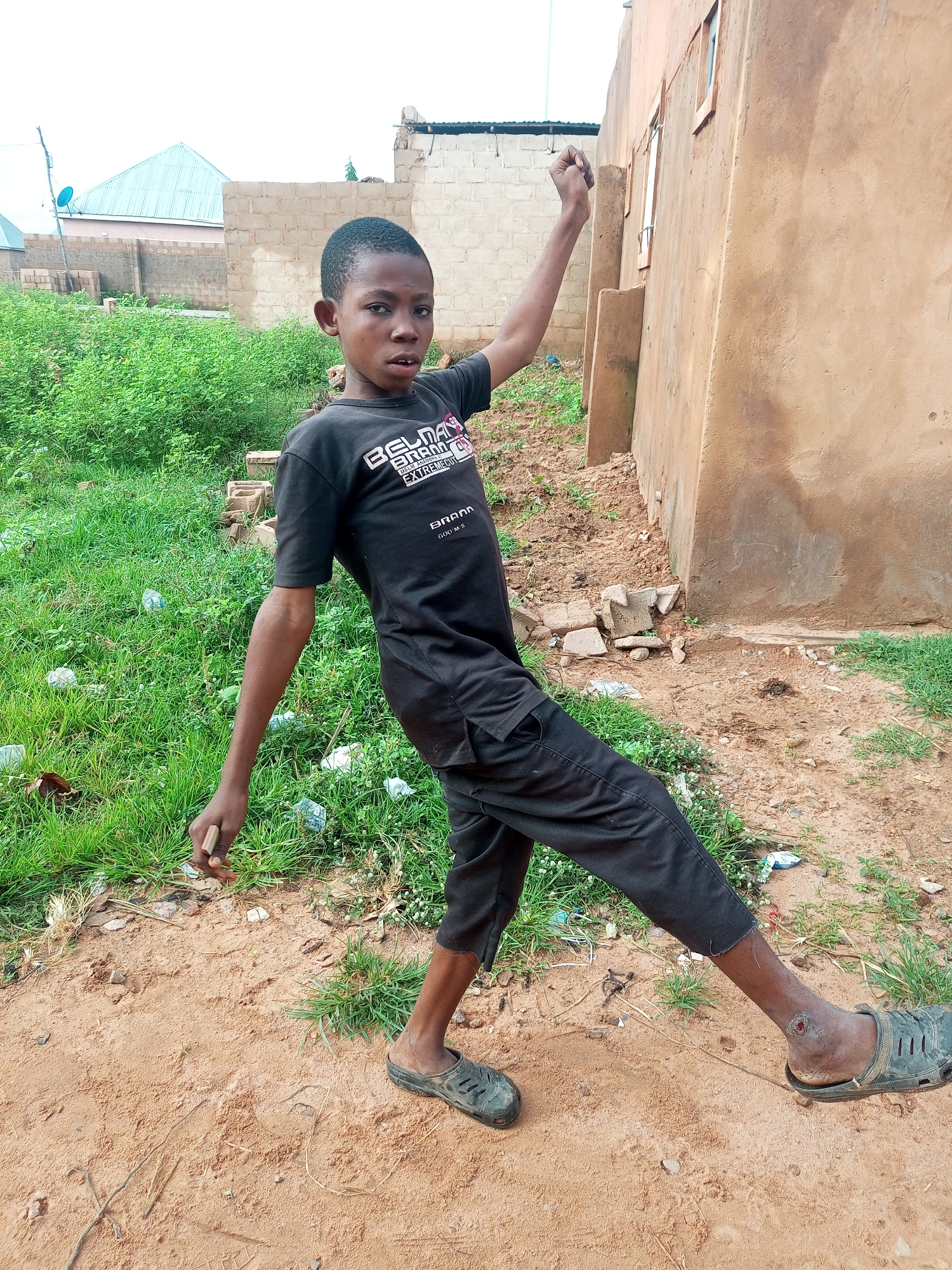 A young boy with dark skin and short, dark hair stands in a semi-squatting position with his right arm raised and his left hand extended downwards. He is wearing a black t-shirt with white lettering and black pants. His stance suggests a playful or energetic pose. He is wearing dark sandals. The ground is covered in sand and dry leaves, with patches of green grass. In the background, a light brown, unfinished building stands. The environment appears to be an outdoor area in a developing part of a city, possibly in Tempure, Nigeria, characterized by its arid ground and simple construction.