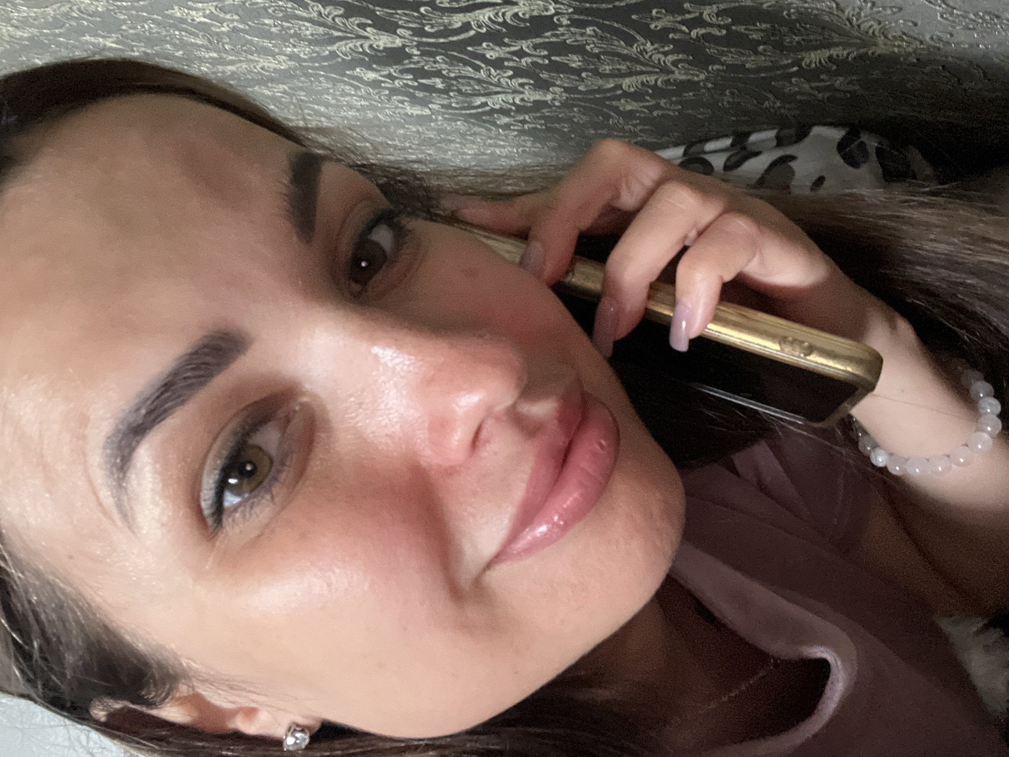 A medium shot shows a woman holding a golden cell phone to her right ear, with her face turned slightly towards the camera. Her left hand is visible, supporting the phone, and her nails are painted a light pink. She is wearing a pink top and a bracelet made of small white beads on her left wrist. The background consists of a patterned wallpaper with a damask design in shades of grey and gold. The lighting appears to be natural, highlighting the woman's features, including her brown eyes and glossy pink lips.