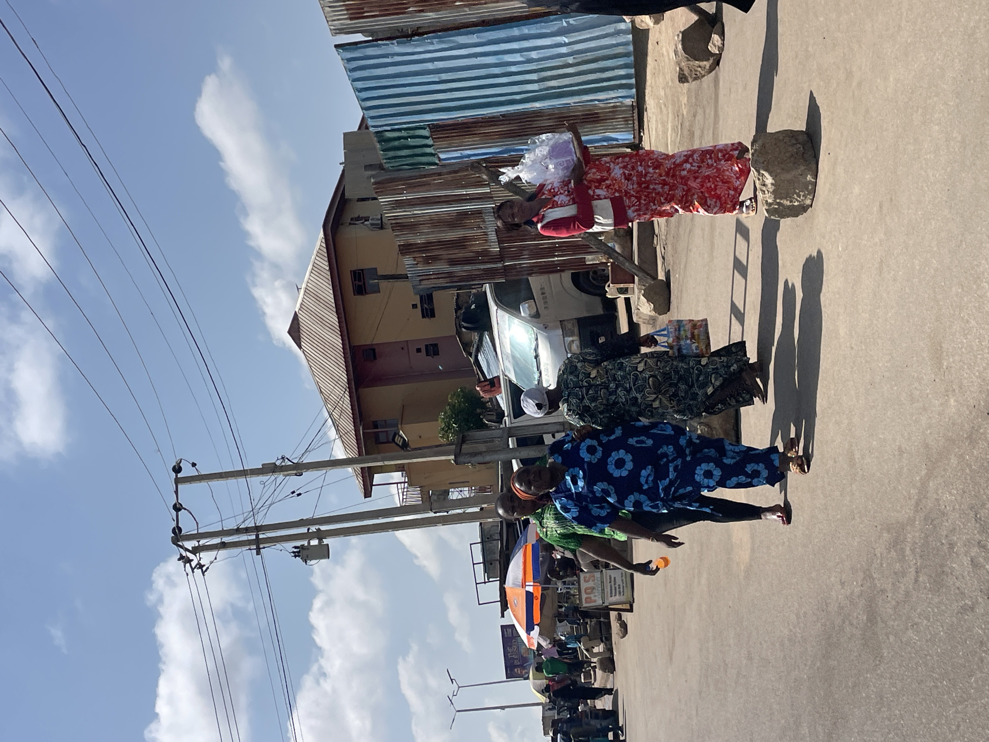 In Jos, Nigeria, a street scene unfolds under a bright, cloudy sky. A group of people walk on a paved road. In the foreground, a woman in a vibrant red and white floral dress carries a clear plastic bag filled with purple items, balanced on a tray. She wears a white vest over her dress and white sandals. To her left, another woman in a blue and white floral dress walks, her left arm extended. Behind them, a man in a green and black patterned shirt walks with his left arm extended. Further left, a man in a blue patterned shirt and dark pants walks with a bottle in his hand.
In the background, a white van is parked. A multi-story building with an orange-tiled roof stands behind a corrugated metal fence. Power lines and poles are visible against the sky. An orange and white umbrella shades a market stall. Other market stalls and people are scattered throughout the scene, suggesting a bustling commercial area. Rocks are placed on the road.