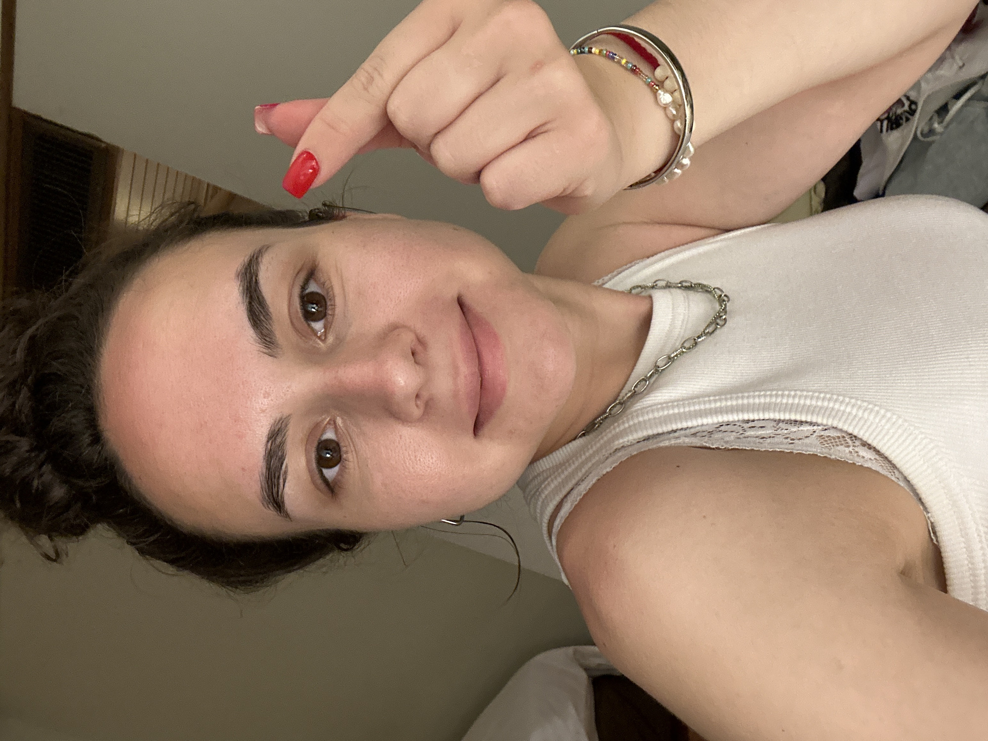 A young woman is posing for a selfie indoors. She is wearing a white tank top with lace trim, a silver chain necklace, and several bracelets on her left wrist. She has dark hair styled in a messy bun and brown eyes. Her left hand is raised, with the index finger and thumb forming a small heart shape. Her fingernails are painted red. The background is blurry but appears to be a neutral-toned interior. The photograph was likely taken in a hotel or residence. There is no information to confirm the location is in Pattaya City, Thailand.