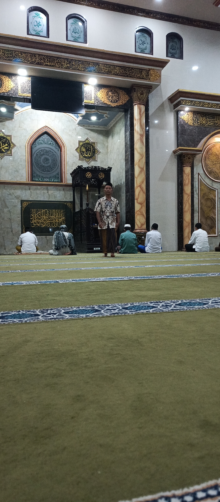 The image shows the interior of a mosque in Arjasa, Indonesia. The mosque features traditional Islamic architecture with ornate decorations and calligraphy on the walls. The floor is covered with a green carpet with blue and white patterned stripes, indicating prayer rows. Several men are present, some are seated on the carpet facing towards the front of the mosque, likely engaged in prayer or waiting for prayer to commence. One man stands in the foreground, facing the camera, dressed in a patterned short-sleeved shirt and a sarong. Another man stands near a decorative wooden pulpit. The lighting in the mosque appears to be artificial. The overall atmosphere suggests a place of worship during a service or a gathering.