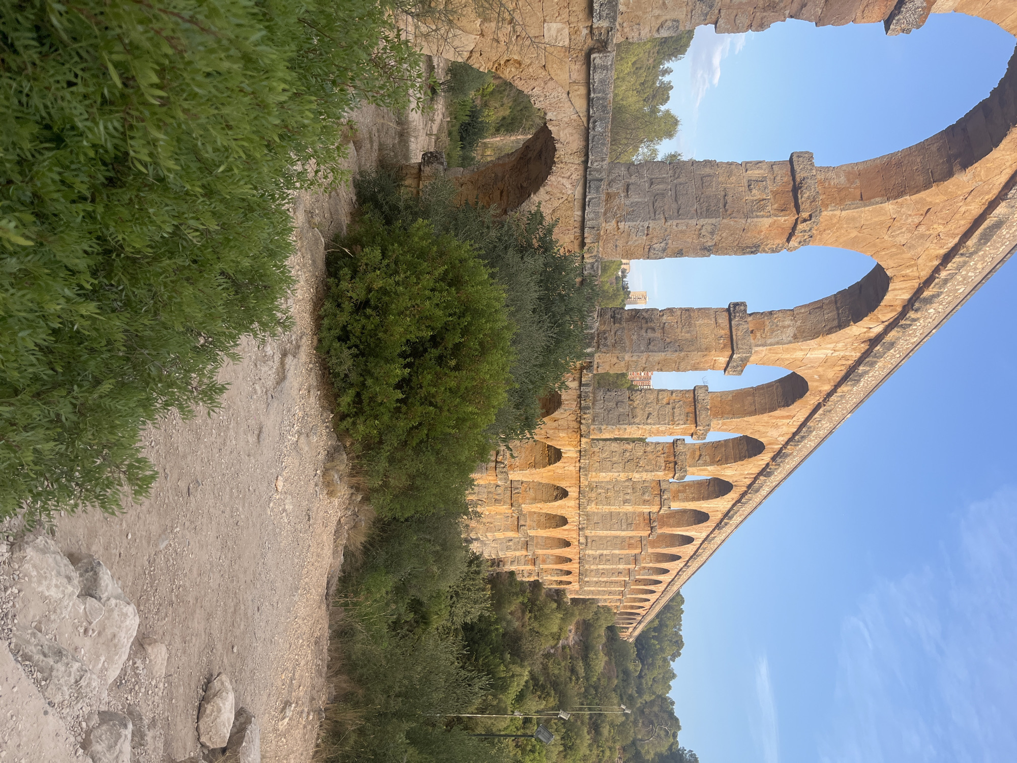 The image shows a large, ancient Roman aqueduct, the Pont del Diable (Devil's Bridge), situated in a rural, hilly landscape on the outskirts of Tarragona, Spain. The aqueduct, constructed of large, reddish-brown stones, features multiple tiers of arches that stretch across the frame, disappearing into the distance. The lower section of the aqueduct has larger arches, while the upper section has smaller, more numerous arches.
The aqueduct is surrounded by lush, green vegetation, including dense bushes and trees that grow on the slopes of the hills. A dirt path, scattered with rocks and pebbles, leads through the foreground and under the aqueduct's arches. The sky above is a clear, bright blue with a few wispy clouds. In the background, a modern building, possibly an apartment complex, is visible in the distance, contrasting with the ancient structure. The scene is bathed in warm, natural light, suggesting it was taken during the late afternoon or early morning. There are no people or animals visible in the image. The overall impression is one of historical grandeur set within a natural environment.