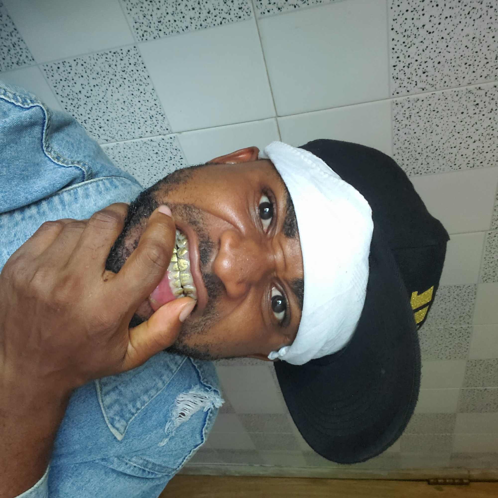 A man with dark skin and a beard is visible in an indoor environment. He is wearing a light blue denim jacket with a torn section. He has a black baseball cap with a white band. He has a full set of gold teeth. The background features a tile ceiling. The photo was taken in Obosi, Nigeria.