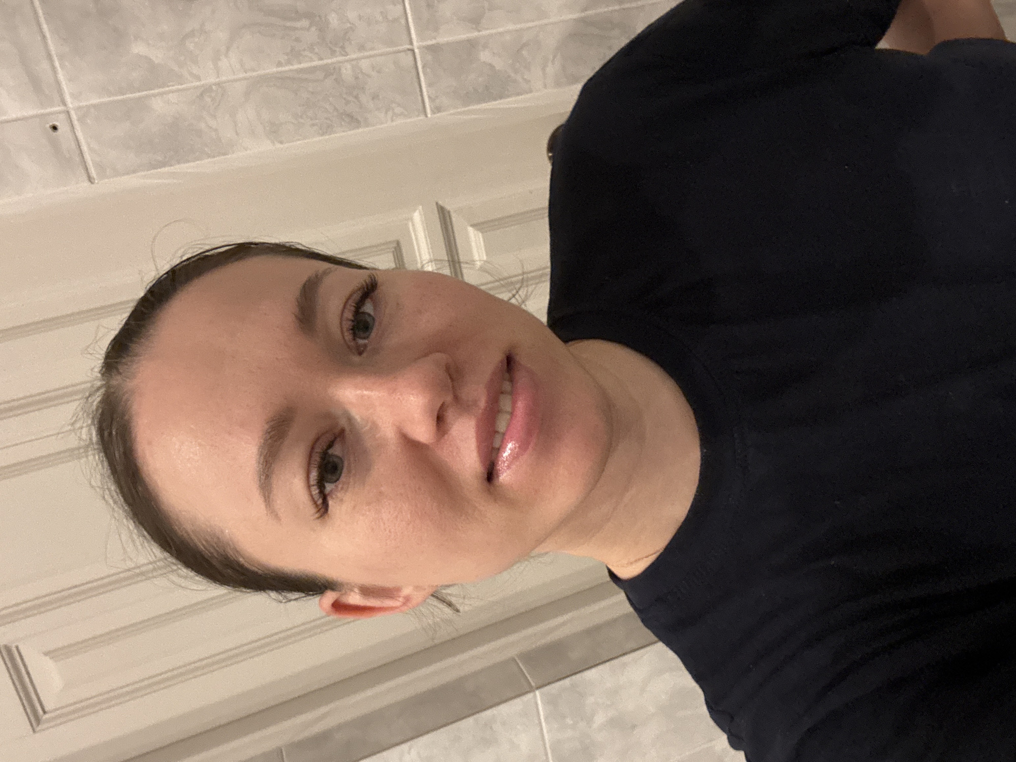 The image shows a woman wearing a black t-shirt. She is indoors in a bathroom, based on the tiled wall and cabinet behind her. The background is out of focus. The location is unknown, and Ферма, Russia, cannot be confirmed from the image.