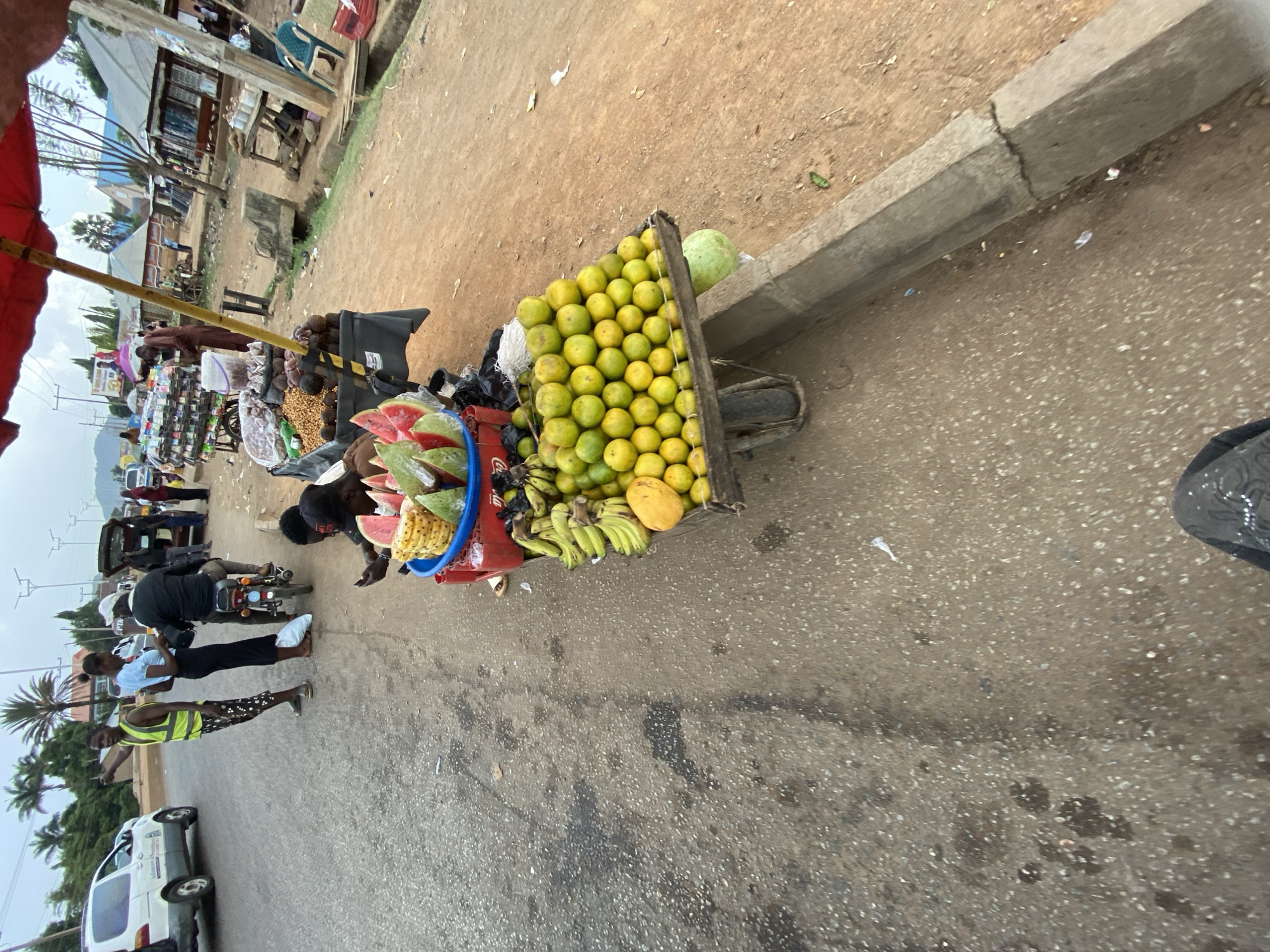 This image captures a bustling roadside scene in Bukuru, Nigeria, centered around a vibrant fruit vendor's stall. The photograph is taken from a slightly elevated perspective, looking across a paved road towards a dirt shoulder and buildings.
In the foreground, a wooden cart is heavily laden with fresh produce. A large mound of green and yellow citrus fruits, likely oranges or limes, occupies a prominent position. Alongside these, bundles of green bananas and a large yellow mango are displayed. A blue plastic bucket holds neatly arranged slices of watermelon and chunks of what appears to be pineapple, ready for purchase. A whole, large green watermelon rests near the oranges. Various other items, possibly bagged snacks or nuts, are also visible on the cart.
Several people are engaged in activity around the stall and on the street. A vendor, dressed in dark clothing, attends to the fruit display, while a customer leans over the cart, seemingly selecting items. Further down the road, a person wearing a bright yellow safety vest stands near a white van, pointing towards the left. A motorcycle with a rider is stopped on the road, and other individuals are seen walking or standing in the background, contributing to the busy atmosphere.
The setting includes an asphalt road, a concrete curb, and a dirt shoulder. In the background, simple buildings with corrugated iron roofs and some sparse vegetation are visible. The lighting suggests it is daytime, possibly under an overcast sky, as shadows are soft. A red umbrella canopy is partially visible in the upper left, providing shade. On a red crate supporting the fruit stall, the brand "Coca-Cola" is clearly discernible. The scene illustrates everyday commerce and life in a Nigerian community.