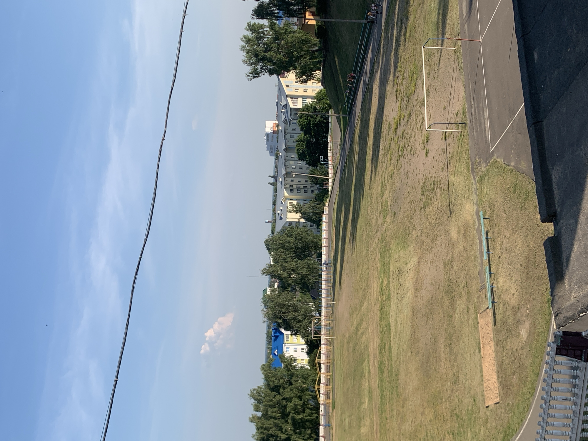 The image shows an outdoor scene on a clear day, likely in the late afternoon given the long shadows cast across the grass. The primary subjects are a grassy field that appears to be part of a sports complex, with a white soccer goal visible in the middle ground. Adjacent to the field is a paved track with white lines. In the background, several multi-story apartment buildings with light-colored facades and some with blue roofs are nestled among lush green trees. A power line stretches across the top left portion of the frame, with a few distant, small dark specks that could be birds or distant aircraft. The overall impression is one of a calm, suburban or urban park setting. There is no visible text in the image. The location context of Kapyĺ, Belarus, suggests this is a view of a residential area with recreational facilities.