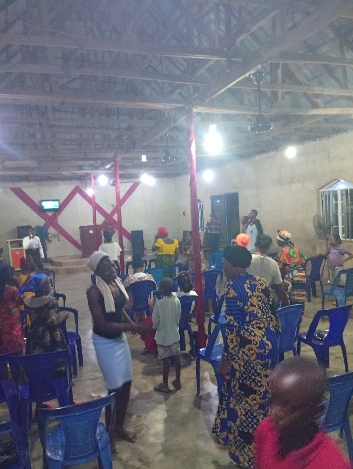 Inside a church with blue chairs and a wooden ceiling, people gather. A woman in a dark top and light skirt stands barefoot, while another wears a blue floral dress. Children are scattered among the adults. A stage decorated with red shapes is visible in the background, and a speaker system stands nearby. The room is illuminated by several bright, bare bulbs hanging from the ceiling.