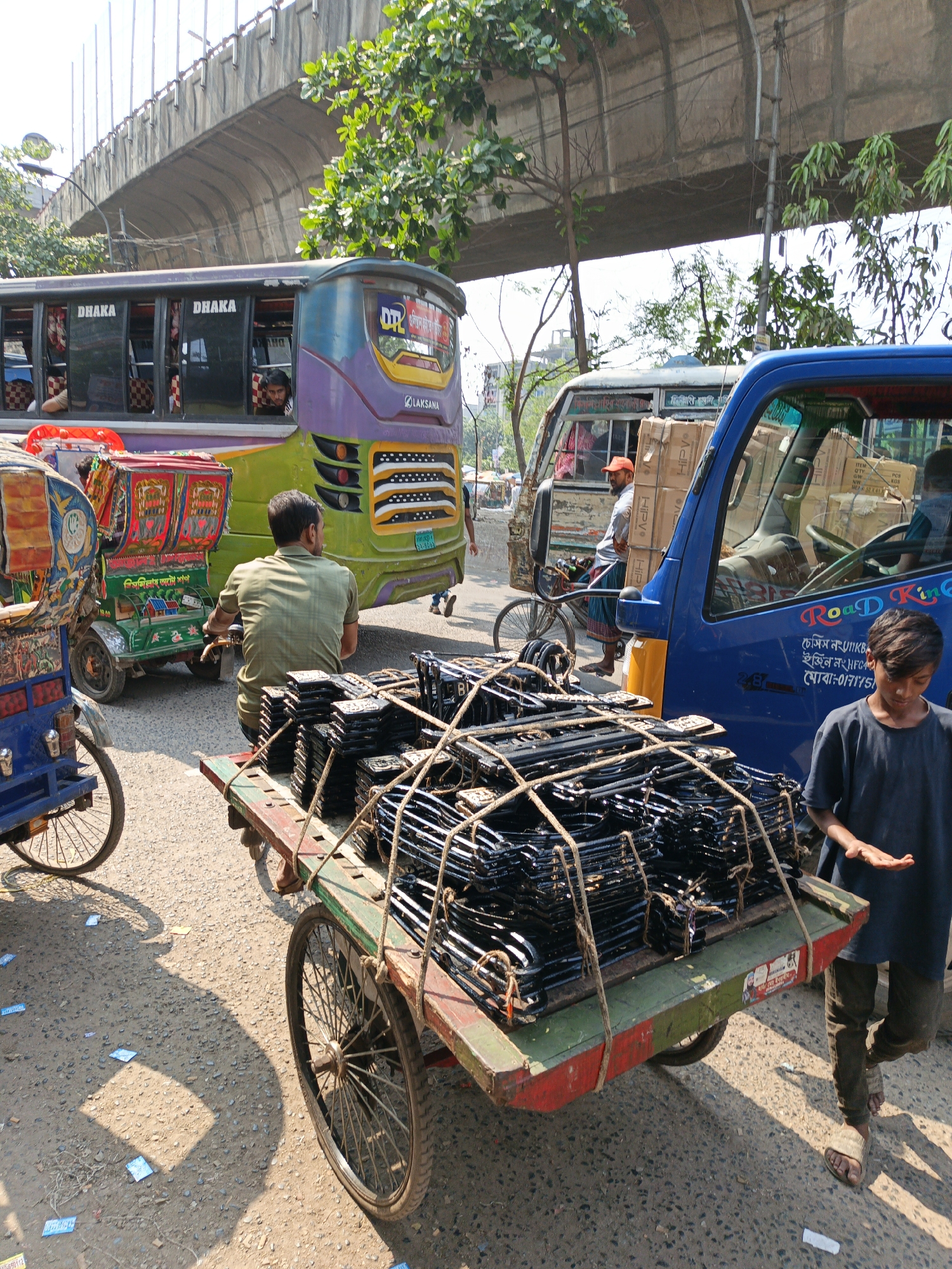 The image captures a bustling street scene in Dhaka, Bangladesh, characterized by a mix of public transportation, informal commerce, and pedestrian activity. In the foreground, a three-wheeled cart, heavily loaded with stacks of black metal components tied with rope, is being pulled along. A barefoot young man in a dark blue shirt walks alongside the cart, gesturing with an open hand, possibly seeking alms or offering assistance.
Behind the cart, a man in a light green shirt sits on a similar three-wheeled rickshaw, facing away from the camera, while another individual operates a vibrant, painted rickshaw. A large, green and purple bus with the word "DHAKA" displayed prominently in its windows is partially visible, with passengers seen looking out. To the right, a small blue truck carrying cardboard boxes is present. A man wearing an orange cap and a sarong stands by it.
The scene is set on a paved road with litter scattered around. Above the street, a concrete overpass arches, adding to the urban density of the location. The presence of trees lining the road suggests a street that is part of the larger Dhaka area, reflecting the typical urban environment of the city. The scene is dynamic, capturing the everyday movement and economic activities of the city's inhabitants.