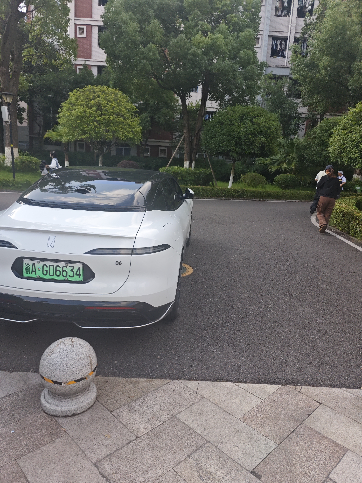 A white electric car with a green license plate that reads "渝 A G06634" is parked on an asphalt surface in front of a building. The license plate is from Chongqing. The car has a black roof and tinted windows. Behind the car, a building with red and white brickwork is visible, surrounded by lush green trees. Two people are walking away from the camera on the right side of the image. One person is wearing a dark jacket and brown pants, while the other person is wearing a white shirt and a hat. A gray stone bollard with yellow and black stripes is in the foreground, on a paved area with stone tiles. The environment appears to be a residential area or a public space within the Chongqing High-tech Zone in China.