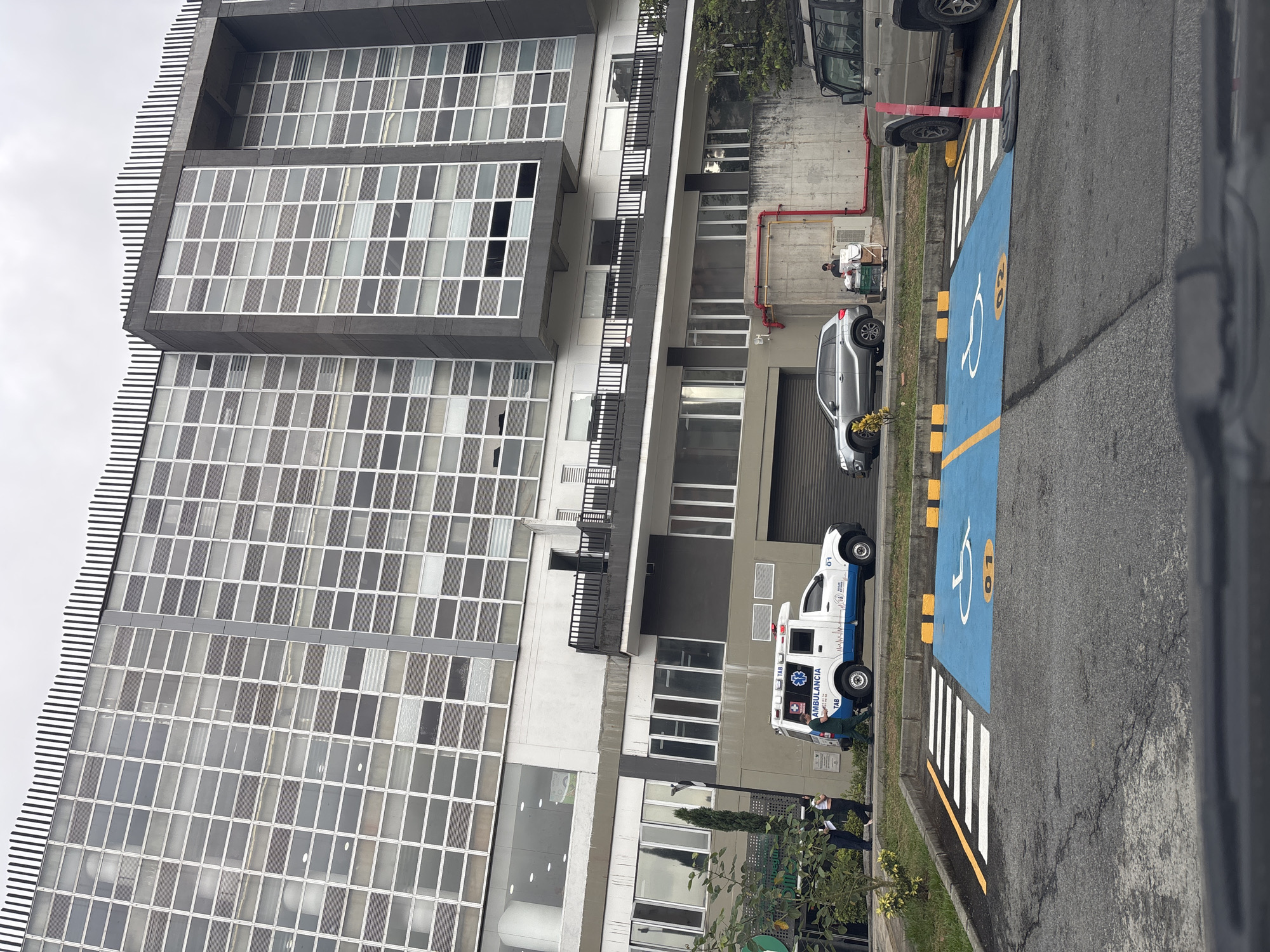 An ambulance is parked next to a handicap parking spot in front of a modern building in Bello, Colombia. The building has a glass facade and horizontal stripes at the top. A silver SUV is parked nearby. There is an individual sitting by a dolly with some items on it further down the street. There are two people walking on the sidewalk near the ambulance.