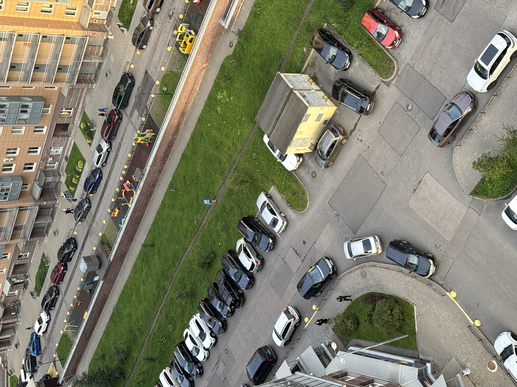 An aerial perspective captures a residential cityscape in an unknown location in Russia, dominated by a large, multi-story apartment building with beige and brown brickwork. The building features numerous balconies with glass railings and exposed concrete elements. In front of the building, a paved courtyard area is visible, lined with parked cars. These vehicles are predominantly sedans and SUVs, with colors ranging from black, silver, white, and red. A children's playground with colorful structures and swings is situated adjacent to the courtyard. A strip of well-maintained green grass separates the courtyard from a network of pathways and additional parking areas. Several individuals are present in the scene, including two people walking in the courtyard and one person walking on a pathway. A small, yellow transformer box with a dark roof is also visible in the foreground. The environment appears to be urban, with a focus on residential infrastructure and recreational spaces.