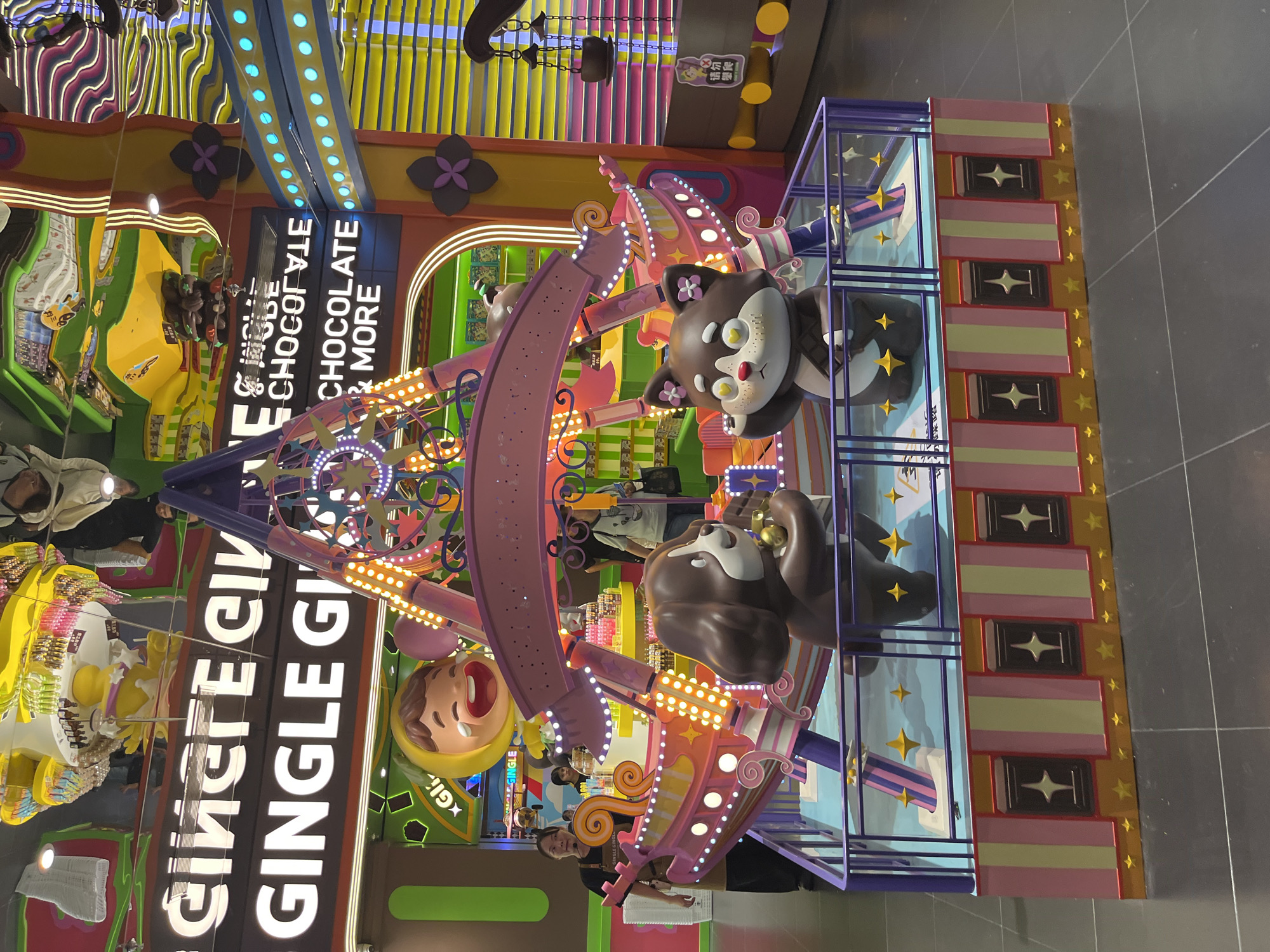 The image shows a brightly decorated indoor amusement ride, likely a carousel or miniature Ferris wheel, located in a commercial space. The ride features cartoonish figures of a dog and a cat. In the background, a sign displays white text, partially obscured and reversed due to reflection, that appears to read "GINGE GINGE" and below it "CHOCOLATE & MORE". The environment is characterized by vibrant colors, flashing lights, and reflective surfaces, suggesting a playful and engaging atmosphere. Several people are visible in the background, appearing to be shoppers or staff members. One woman in the foreground, wearing an apron and headset, is facing the ride. The presence of a sign with Chinese characters, though partially illegible in the reflection, and the overall aesthetic suggest this location is likely a themed retail or entertainment establishment within Haizhu District, China.
