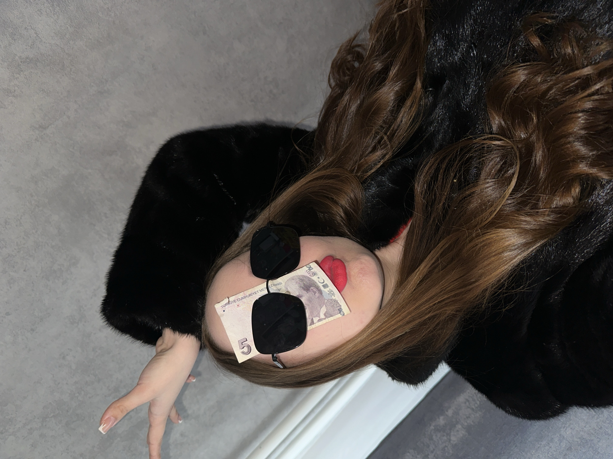 A person is lying down wearing a fur coat and sunglasses. A 5 Turkish lira note is placed under the sunglasses. The person has red lipstick and light brown hair. The background is a gray wall and a white baseboard. It is not possible to confirm the location in the city of Chaikovsky, Russia, from the image alone.