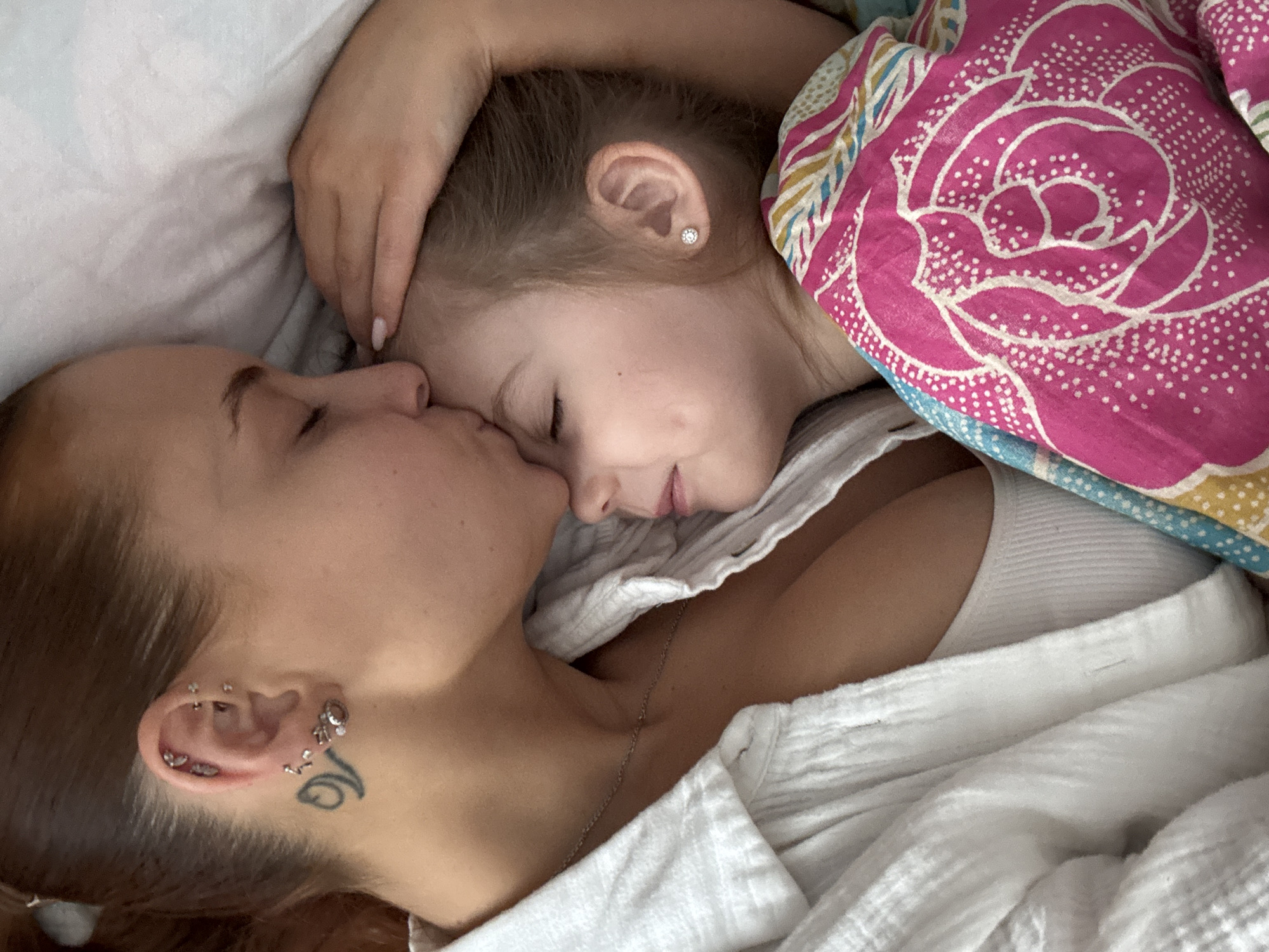 A woman and a young girl are lying in bed, with the woman kissing the girl on the nose. The girl is partially covered by a blanket with a pink floral pattern. The woman has multiple piercings in her ear and a tattoo near her ear. They are lying on a white sheet in the city of Chastyye, Russia.