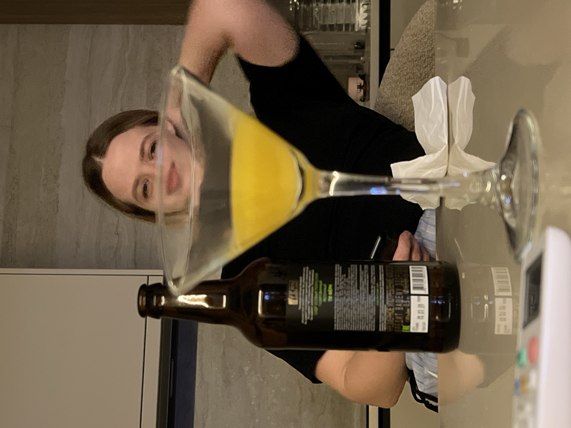 A woman in a black shirt is holding a glass of liquid with a brown bottle in front of it. There are white napkins and a white remote on the table. Based on the limited visual information available in this image, it is not possible to determine the precise location within the city of Рубцово, Russia.