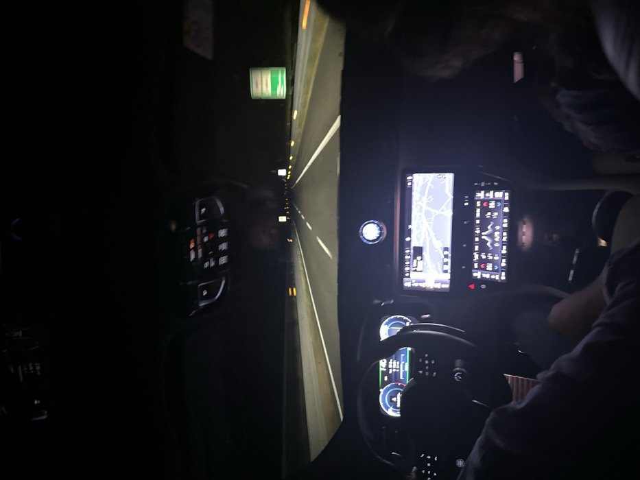 Inside a vehicle at night, illuminated displays showcase a digital map and car metrics. A steering wheel and dashboard are visible. Outside, a highway stretches ahead, marked by white lines and illuminated by intermittent lights. A green sign is seen on the roadside. The scene is dark, with the car's interior lights providing the main source of illumination.
