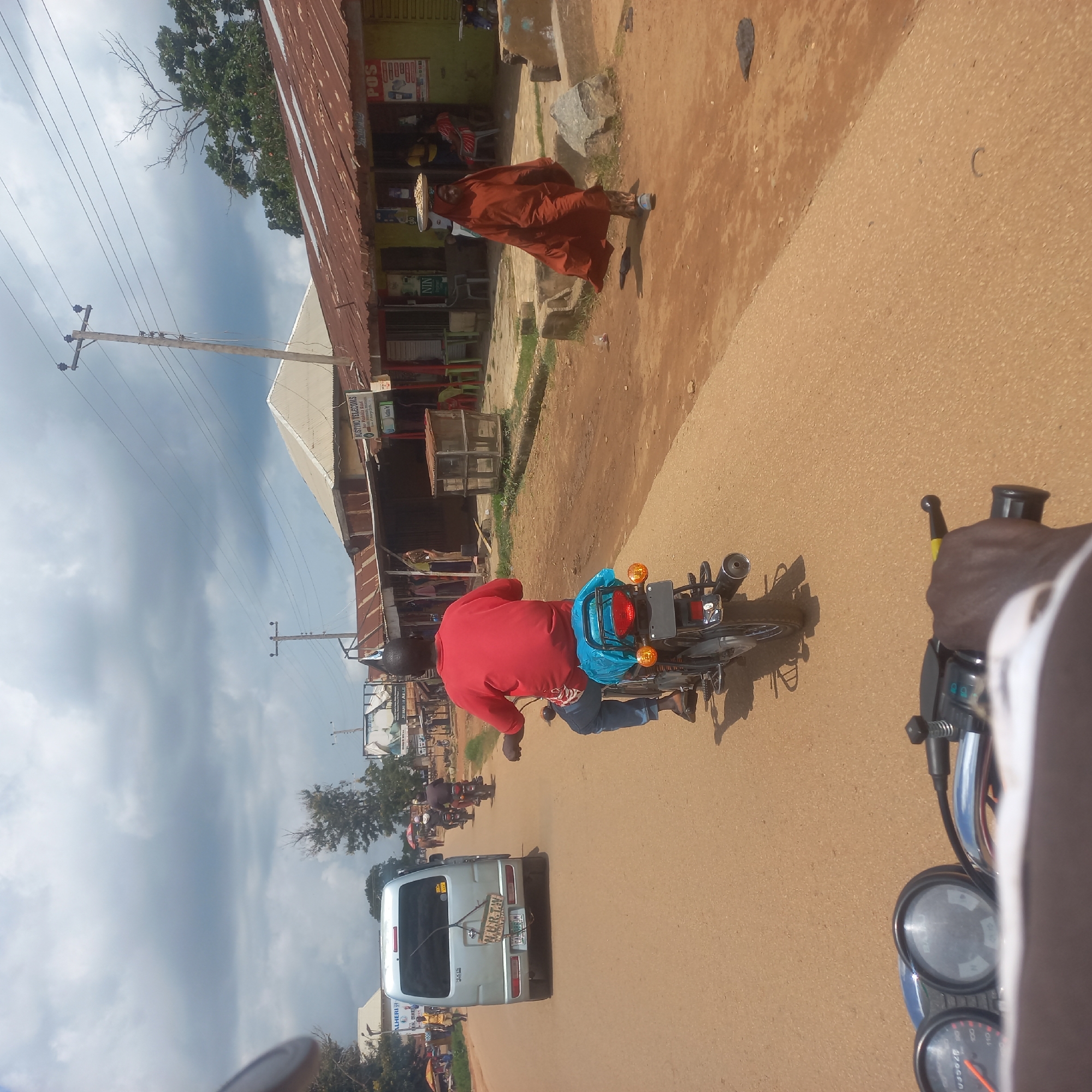 In Mangu Town, Nigeria, under a cloudy sky, a person in a red shirt and blue jeans rides a motorcycle on a paved road. The motorcycle has a blue plastic bag covering its rear seat. Further down the road, a silver van is parked. On the right side of the road, several small shops with corrugated metal roofs are visible. One shop has a sign that says "POS." A woman wearing a burnt orange headscarf and covering her body walks on the dirt path beside the road, carrying a tray of items on her head. Other motorcycles and people are seen in the background, suggesting a bustling street. The environment appears to be a typical commercial area in a Nigerian town, with a mix of businesses and traffic.