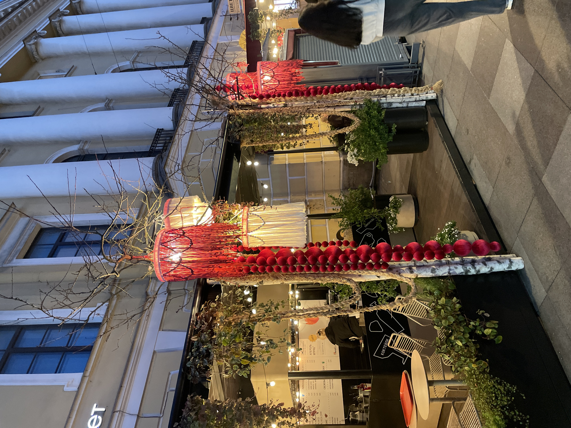 This evening street view in Saint Petersburg, Russia, showcases the elaborately decorated storefront of a commercial establishment, set against the backdrop of a building with classical architecture. The upper floors of the building feature distinctive tall, white columns and arched windows, suggesting a historical or grand structure.
The ground-level storefront is adorned with a festive display. Two large, cylindrical light fixtures, one in vibrant red and another in white, hang prominently, casting a warm glow. These lights are integrated into a vertical arrangement of bare tree branches, giving a rustic yet artistic feel. Below the main lights, rows of red spherical ornaments, resembling garlands, are strung across the facade. Additional decorations include braided ropes and potted greenery, enhancing the inviting atmosphere. Small string lights are subtly woven throughout the display.
On the tiled pavement outside, a person is partially visible, walking away from the storefront. Through the glass entrance, dimly lit interiors reveal menu boards, tables, and chairs, suggesting a cafe or bar.
Visible Cyrillic text includes "ЦВЕТ," which translates to "color" or "blossom," potentially indicating the establishment's name. Other text lists menu items such as "ЧАЙ" (Tea), "ЛИМОНАД" (Lemonade), "СВЕЖЕВЫ" (likely part of "Freshly squeezed juices"), "АПЕРОЛЬ ШПРИЦ" (Aperol Spritz), and "КОКТЕЙЛИ" (Cocktails), with a price of "249 P" (249 Rubles) for some offerings. The scene evokes a welcoming and aesthetically pleasing urban evening.