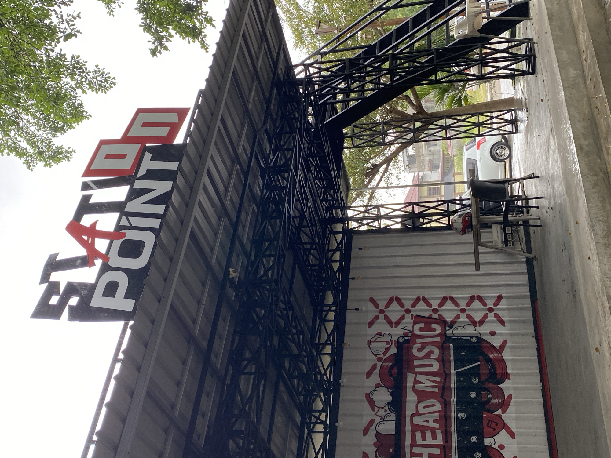 An outdoor scene in Lhokseumawe, Indonesia, captures a modern, industrial-style building under a bright, overcast sky. The image is oriented vertically. A large sign, featuring red and white letters on a dark background, proudly displays "STATI POINT". Below it, a white corrugated metal wall, possibly a rolling shutter or door, is adorned with a bold graphic of a cassette tape and the text "HEAD MUSIC" in red and black.
The building itself incorporates corrugated metal siding and an intricate black metal truss framework, suggesting a contemporary architectural design, potentially for a cafe or recreational spot. The ground beneath the structure appears wet, indicating recent rain or damp conditions. In the background, beyond the metal framework, lush green trees are visible, adding a natural element to the urban setting. A white compact car, possibly a Suzuki Wagon R, is parked on the wet ground, and a black chair along with another lighter-colored chair or stool are placed nearby, suggesting an outdoor seating area. No people are visible, and no specific activity is taking place. The time of day appears to be daytime, given the bright illumination.