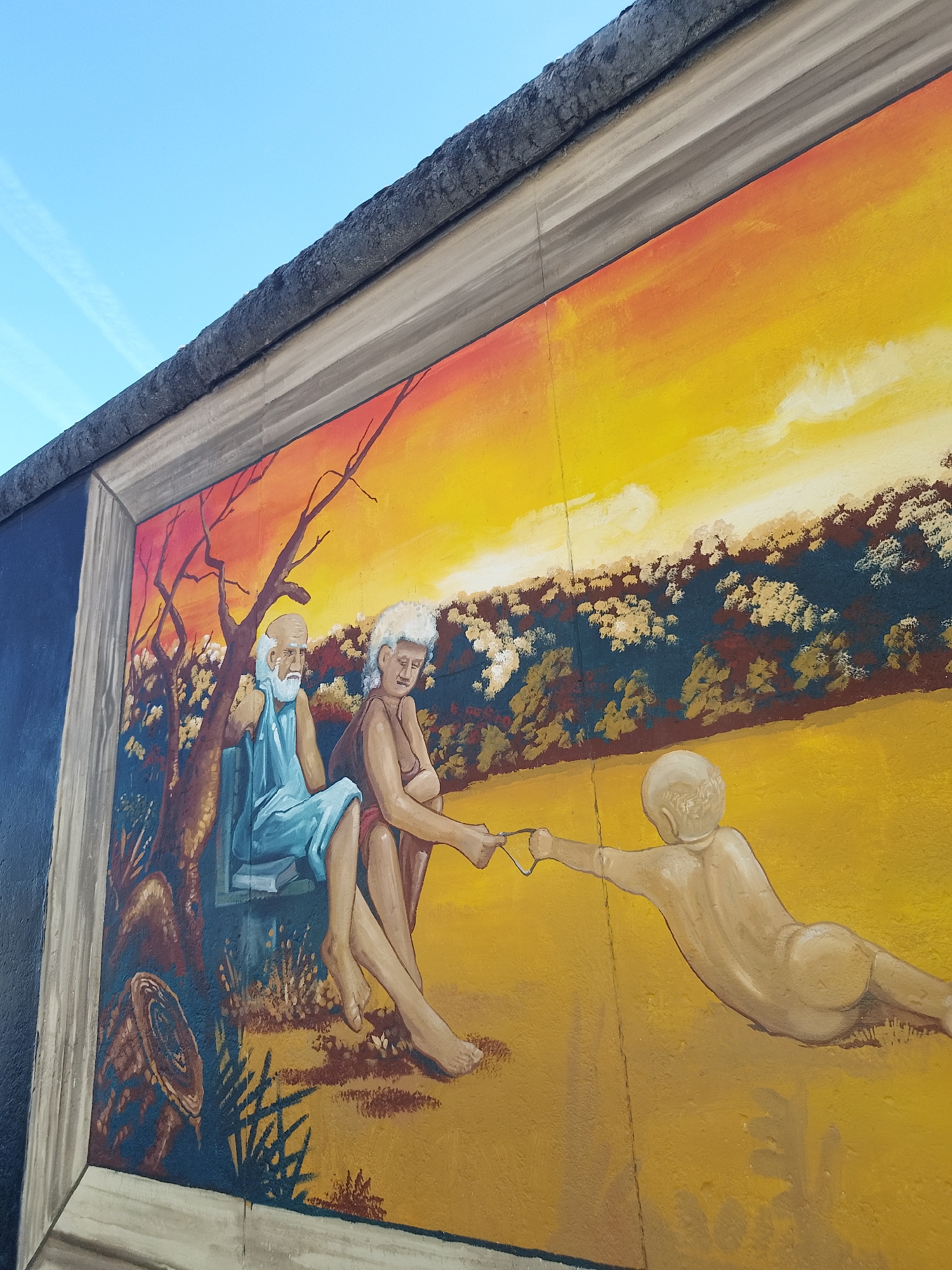 This image shows a section of the East Side Gallery in Berlin, Germany. The mural depicts an elderly couple seated in a landscape, extending a wire or string toward a figure lying prone on a field of yellow. The backdrop features a sunset sky and stylized vegetation. The painting is contained within a painted frame on the wall.