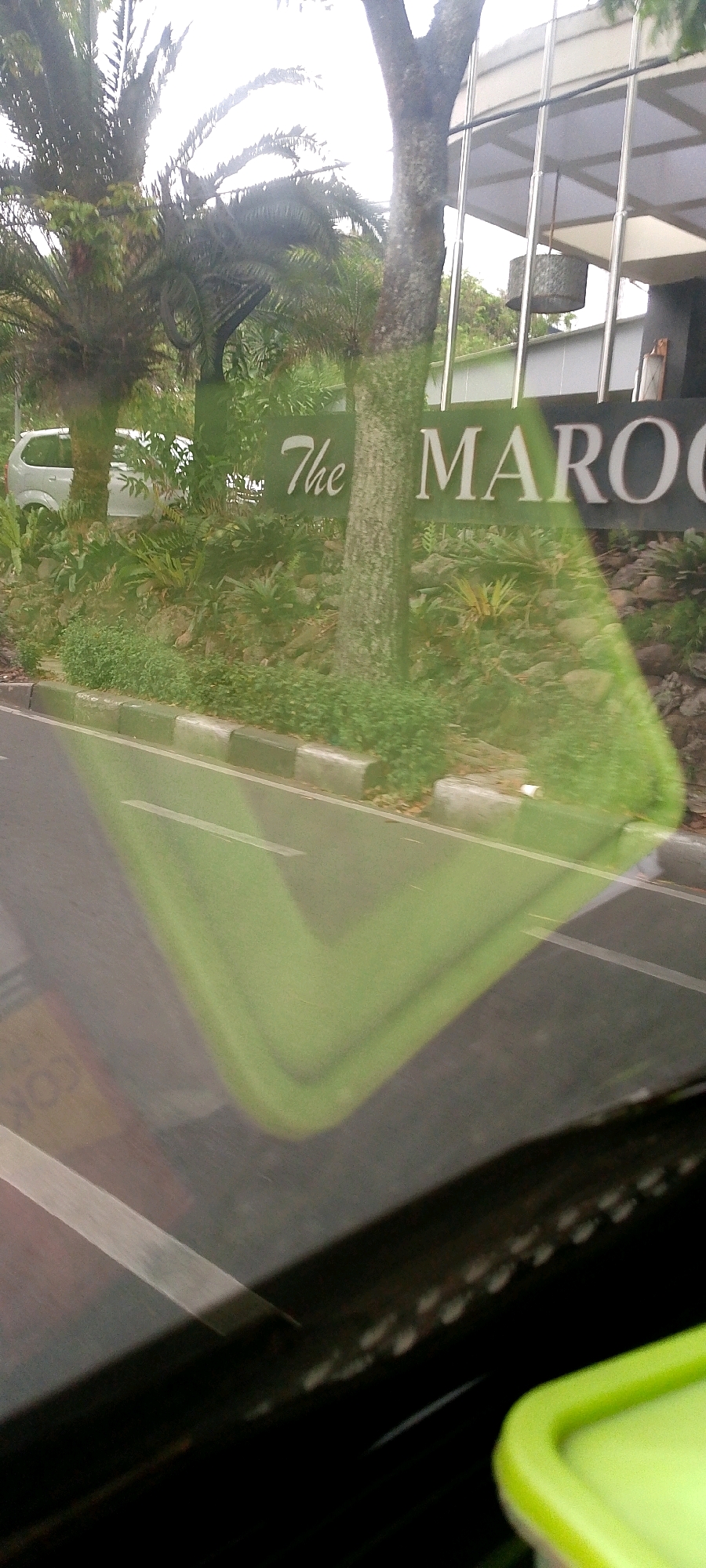 The image shows a sign that reads "The Marocco" on a building with a modern facade. There are palm trees and other greenery in front of the building, and a white car is parked on the side of the road. The image is taken from a car, with a green reflection on the windshield. The scene appears to be in a commercial or residential area.