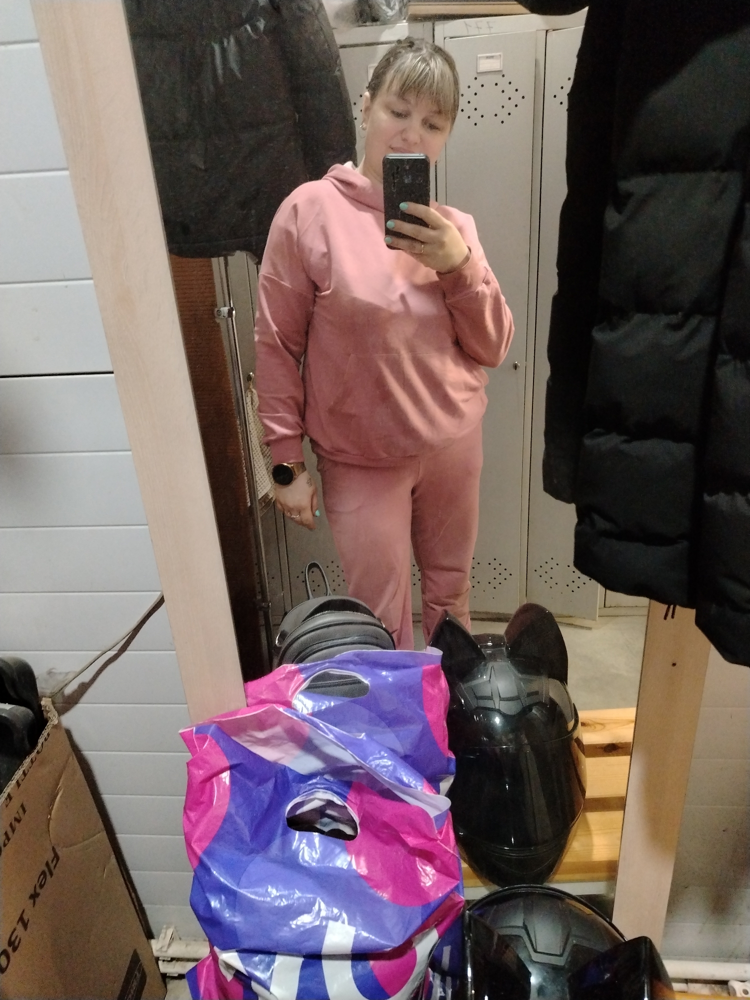 This image captures a mirror selfie taken by a woman in an indoor setting, likely a utility room, garage, or storage area, in Rusanovo, Russia.
The central subject is a woman, reflected in a full-length mirror, taking a photo of herself with a smartphone. She has light-colored hair styled with bangs and is wearing a dusty pink or salmon-colored hooded tracksuit, consisting of a sweatshirt and matching sweatpants. Her nails are painted light blue or turquoise, and she wears a ring on her right hand and a watch on her left wrist. Her expression is neutral as she focuses on her phone screen.
Behind the woman, reflected in the mirror, are two grey metal lockers or cabinets with perforated ventilation patterns, one bearing a small white label. A dark, possibly black, puffer jacket or coat hangs to the right of the reflection. To the left, a light-colored, horizontally paneled wall is visible. A white towel or cloth hangs on a hook behind the woman's left arm.
In the foreground, outside the mirror's reflection, a light-colored wooden mirror frame runs vertically. To the left, a brown cardboard box is visible with partial text that reads "Flex 130" and "IMPA". On the floor or a low surface to the right, a pile of colorful plastic shopping bags, one prominently purple and pink (resembling a Taco Bell bag), is stacked next to two dark, possibly black, motorcycle or bike helmets. One helmet features distinctive cat-ear-like protrusions on top. These items rest on what appears to be wooden planks or a makeshift shelf.
The lighting suggests an indoor environment with artificial light. The overall scene, with the lockers, helmets, boxes, and casual attire, indicates a functional, informal space rather than a living area. The activity is a casual self-portrait.
