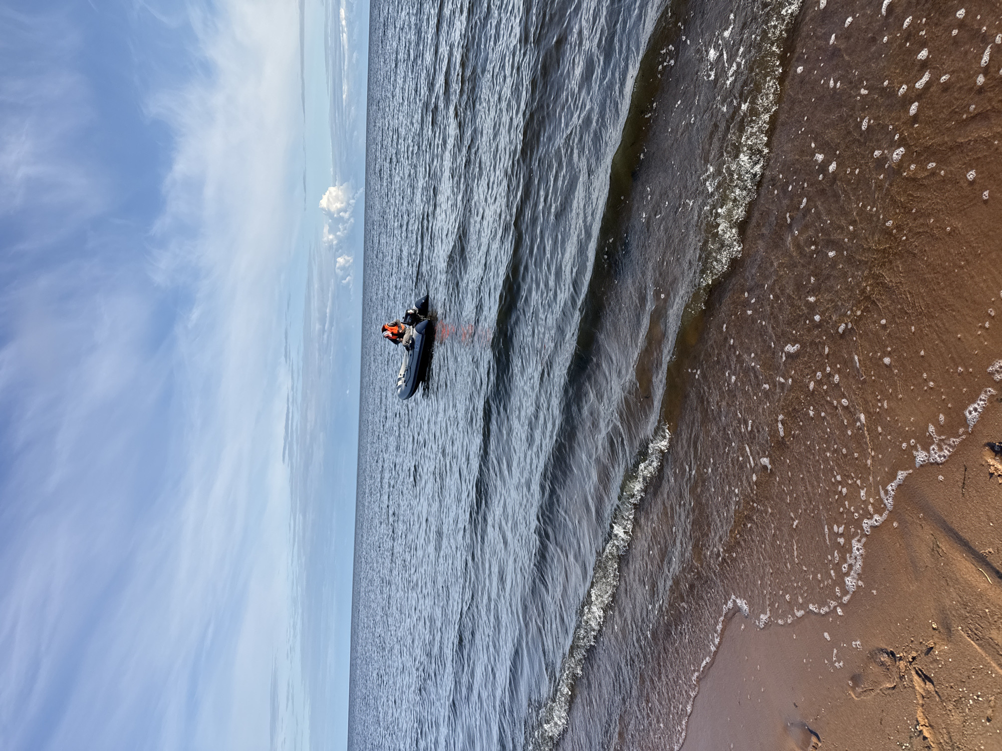 An inflatable boat with a person wearing an orange life vest is on the water. The boat is moving away from the shore. The water is dark blue and calm with small waves. The shore is sandy and brown with some foam from the waves. The sky is blue with white clouds. The scene is likely a beach or coastline.
