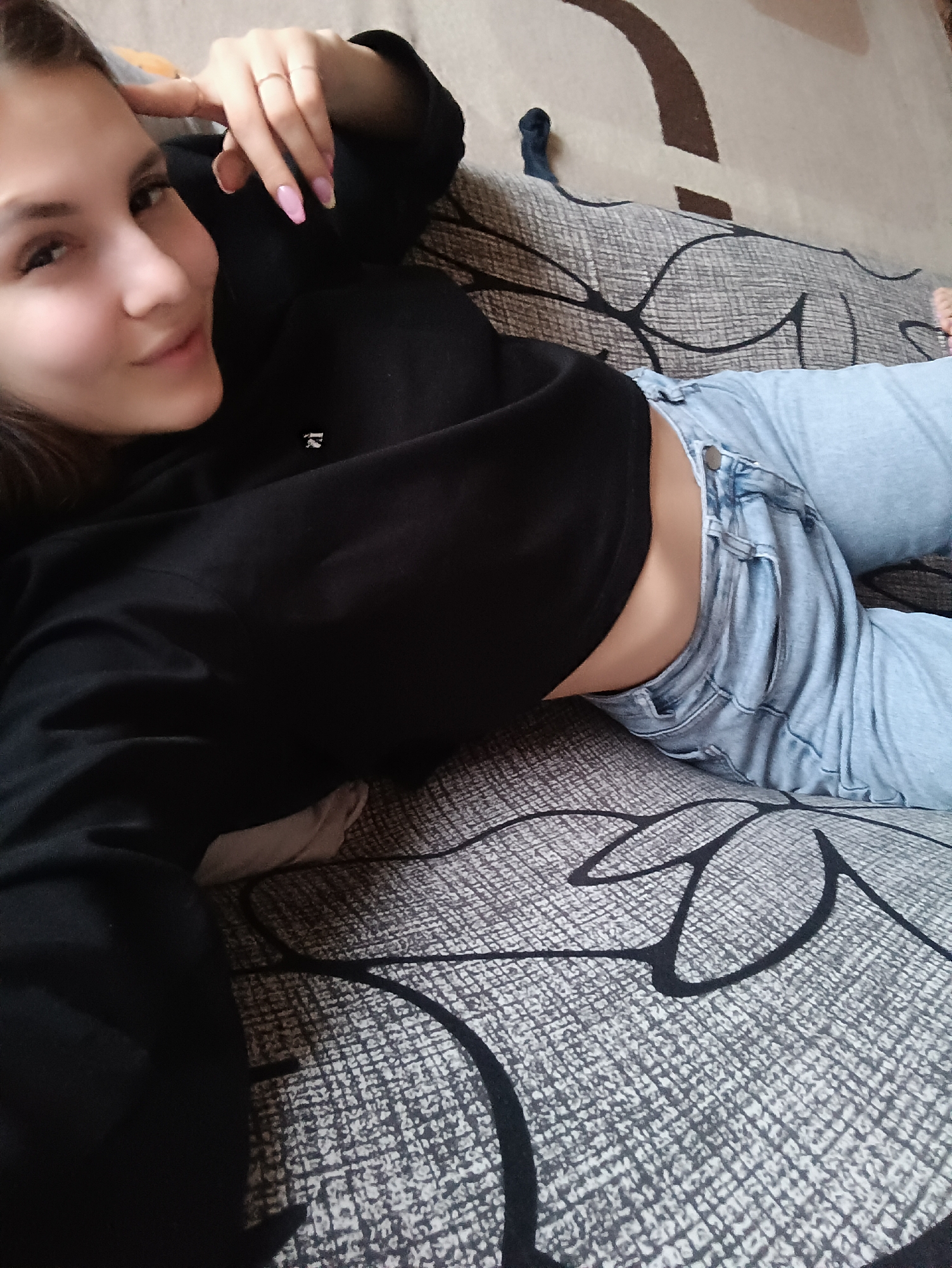A young woman is lying on a patterned gray couch. She's wearing a black cropped sweater and light blue jeans. Her left hand is raised near her head, with rings visible on her fingers. The location may be Chaikovsky, Russia, but that cannot be confirmed from the image.