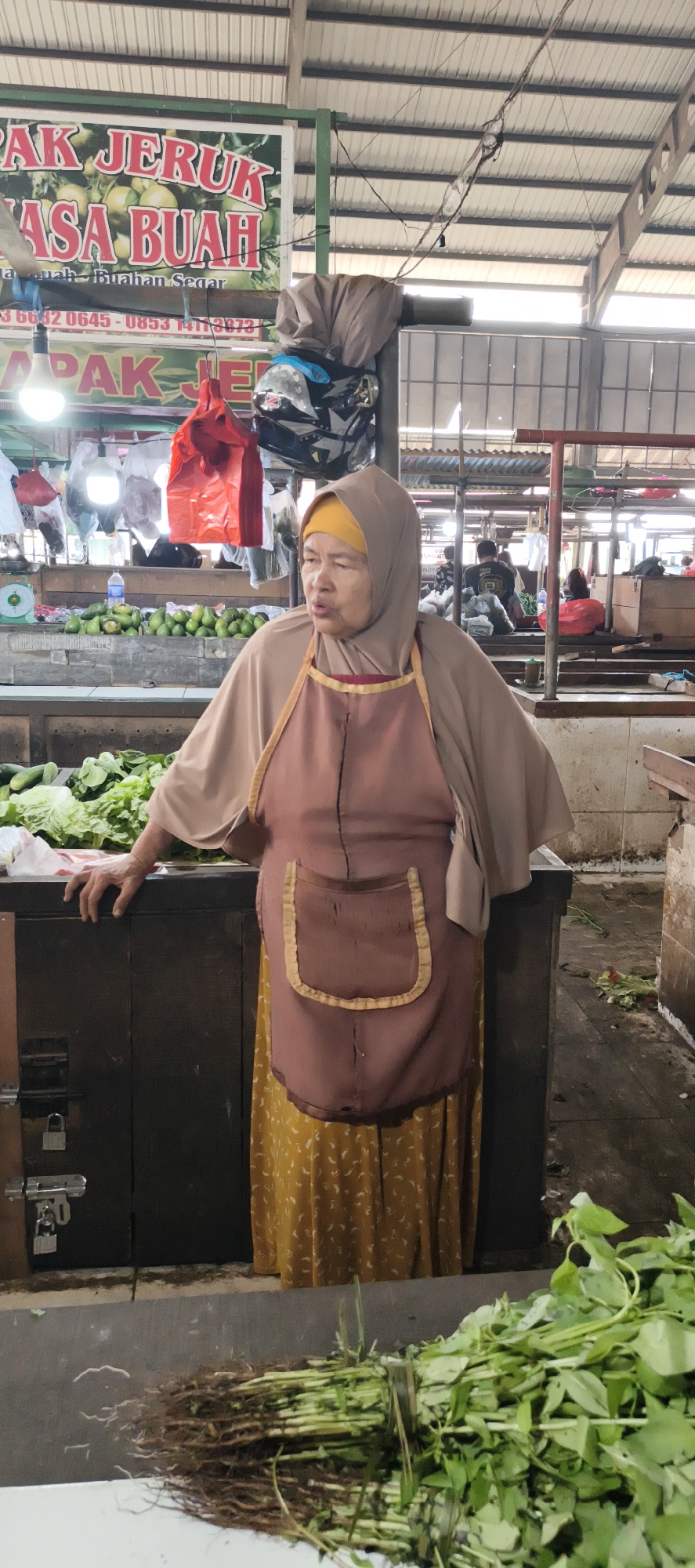 The image captures a street vendor, likely a woman, standing behind a stall piled with fresh produce, primarily leafy greens and what appear to be avocados. She is wearing a light brown hijab and a complementary brown apron over a yellow patterned dress. Her gaze is directed slightly to her right, and her mouth is open as if speaking. The stall is made of dark wood, with a padlocked drawer visible on the left side. Behind her, a sign with Indonesian text, possibly advertising "JERUK" (oranges) and "BUAH" (fruit), is partially visible. In the background, other market-goers and stalls suggest a bustling marketplace environment. The overall setting appears to be an open-air market, typical of Jambi, Indonesia, given the context of the Indonesian language on the sign and the type of produce. The lighting indicates it's daytime, with sunlight filtering through the corrugated metal roof of the market. The presence of hanging bags and the general arrangement of goods are characteristic of a traditional Indonesian market.