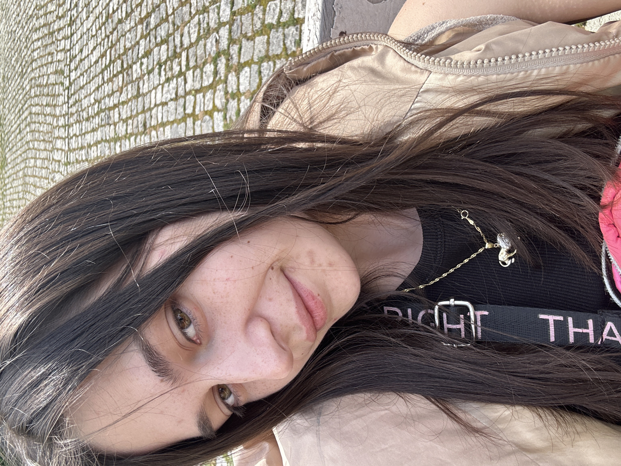 A young woman with long, dark brown hair is the main subject of the image. She has fair skin, light brown eyes, and noticeable freckles on her face. She is wearing a black top with a gold necklace and a tan-colored jacket. A black strap with pink writing is also visible across her chest.
The background appears to be an outdoor setting, likely a sidewalk or paved area, judging from the cobblestone pattern. Based on the information provided, it is possible that the location is in the city of Veliko Tarnovo, Bulgaria, but there are no specific landmarks or details in the image to confirm this. The image is a close-up shot, focusing on the woman's face and upper body.