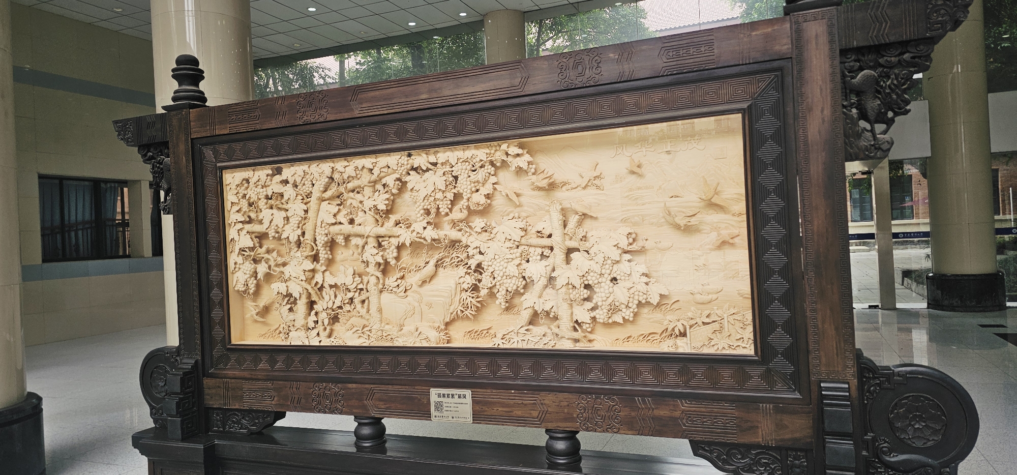 A large, ornately framed wood carving stands in an interior space, likely a lobby or corridor, in the city of Yuzhong District, China. The carving depicts a scene with trees, grape vines laden with grapes, and birds in flight. The frame is dark wood with detailed carvings along its edges and corners. The base of the structure is also dark wood, with curved and sculpted elements. The location appears to be a public building, possibly a cultural center or museum, based on the architecture and the polished tile flooring. A column is visible in the background.