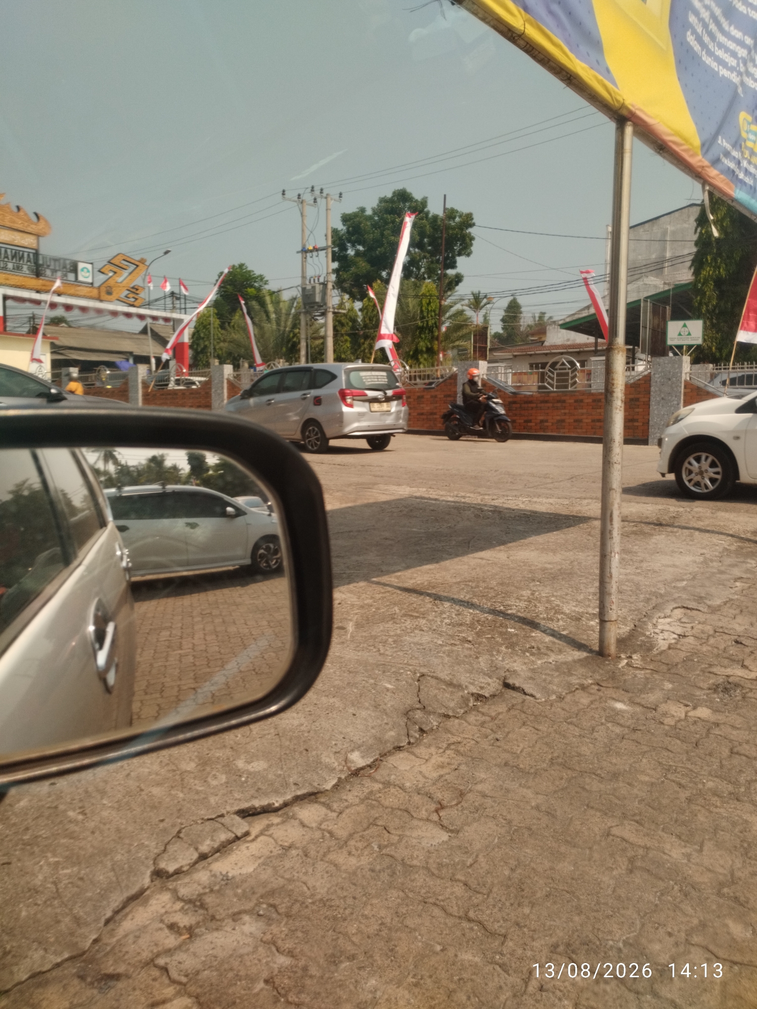 The image captures a street scene in Bandar Lampung, Indonesia, possibly near a school or educational institution, as indicated by the building's architecture and banners. The foreground features the side mirror of a silver car, reflecting another silver car parked on a textured pavement. In the middle ground, a silver minivan is visible, along with a person riding a black motorcycle wearing an orange helmet. The background shows buildings, trees, and utility poles, with several red and white Indonesian flags decorating the scene. The pavement in front of the buildings is made of interlocking bricks, while the road area is concrete with visible cracks. The overall lighting suggests a sunny day, and a timestamp in the lower right corner indicates the image was taken on August 13, 2026, at 14:13.