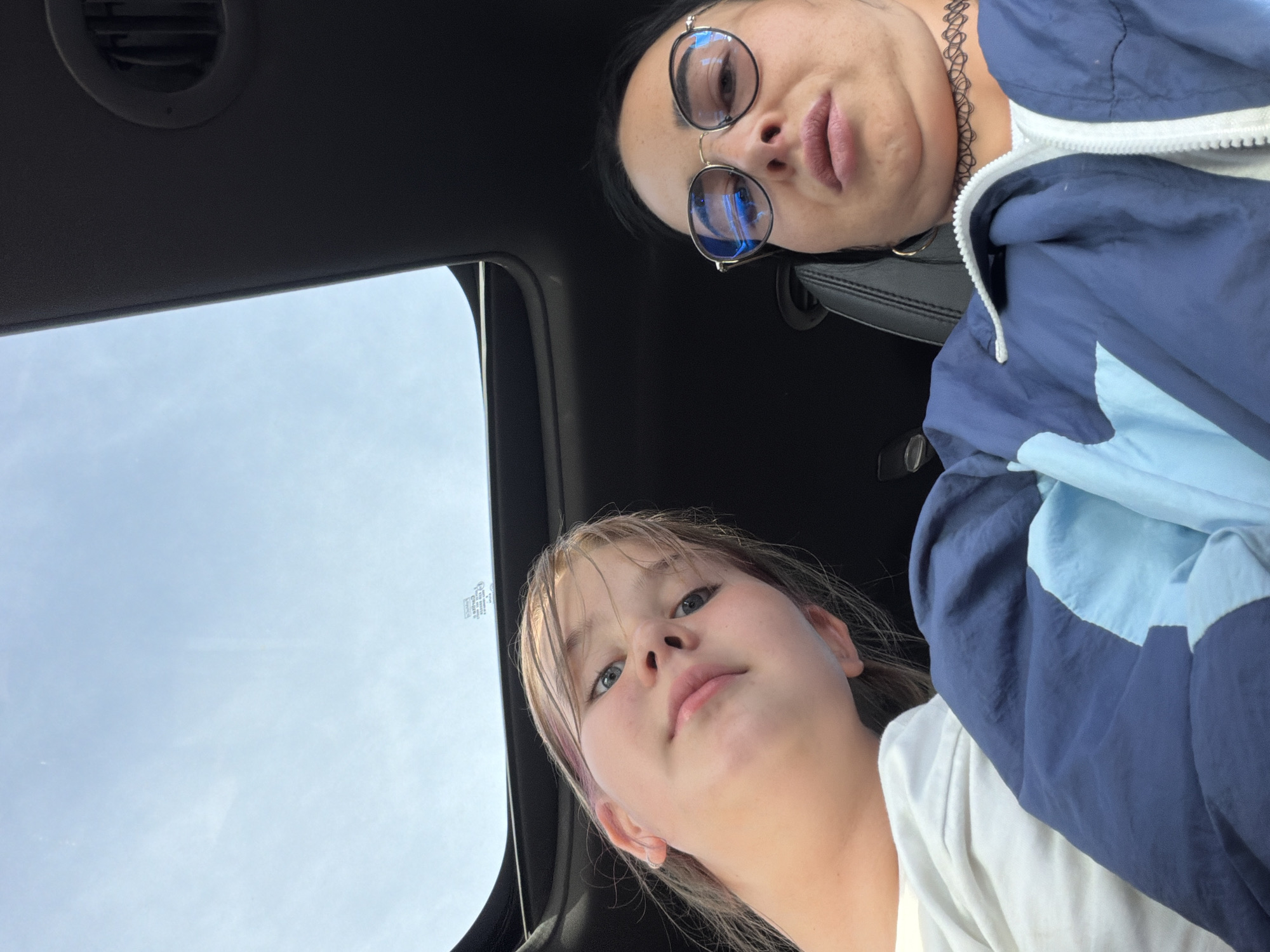 The media file shows a close-up selfie taken from a low angle inside a vehicle, looking upwards towards the sky. Two individuals are visible. In the foreground, a young girl with blonde hair, some streaks of pink, and blue eyes looks directly at the camera with a neutral expression. She is wearing a white t-shirt. Behind her and to the right, an adult woman with dark hair and glasses is also looking towards the camera. She is wearing a blue and light blue jacket and has a choker necklace on. Her expression appears serious. The background shows a sunroof with the sky visible through it, suggesting it is daytime. The scene is likely inside a car with a dark interior, indicated by the presence of a car vent. There are no visible landmarks or specific details that definitively pinpoint the location to Шолохово, Russia, though this context is provided. The weather appears to be clear and bright due to the visible sky.