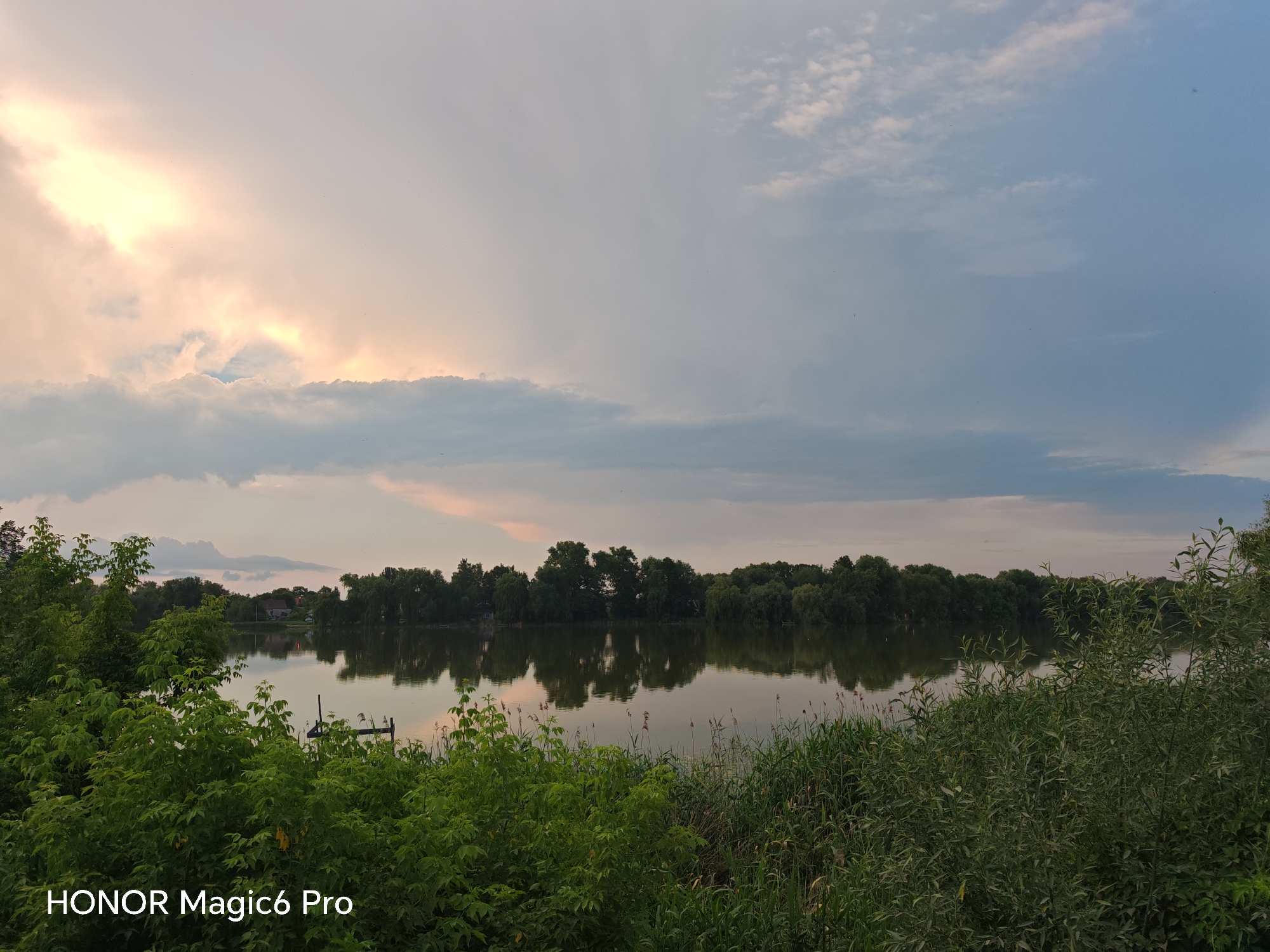 This is a landscape photograph taken in Turbiv, Ukraine. The scene depicts a tranquil lake surrounded by lush greenery and a dense line of trees on the opposite shore. The water is calm, reflecting the trees and the sky. In the foreground, dense foliage and tall reeds frame the view of the lake. A few small, dark objects, possibly boats or buoys, are visible on the water.
The sky is a prominent feature, with dramatic clouds that suggest a transition in weather or time of day. The left side of the sky is illuminated by a bright, diffused light, possibly from the setting or rising sun, casting warm, pastel tones of yellow and pink onto the clouds. The rest of the sky is covered by a softer, grayish-blue cloud cover. A few very small, dark specks, likely birds or insects, are scattered across the sky.
The time of day appears to be either early morning or late evening, given the soft lighting and the atmospheric conditions in the sky. The weather seems to be mild, with a peaceful and serene mood conveyed by the still water and the natural landscape.
In the bottom left corner of the image, the text "HONOR Magic6 Pro" is visible, indicating the device used to capture the photograph. No people are visible in the image. The overall impression is one of natural beauty and a quiet, undisturbed environment.