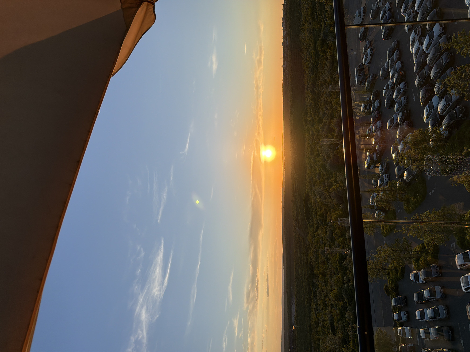 A high-angle shot captures a sunset over a landscape of trees and a parking lot in Stăuceni, Moldova. The sun, a bright orange orb, is low on the horizon, casting a warm glow across the sky and illuminating wispy clouds. The sky transitions from a pale blue at the top to shades of orange and yellow near the horizon. Below the sky, a dense expanse of dark green trees stretches across the scene, with a few sparse buildings visible in the distance. In the foreground, a paved parking lot is filled with numerous cars of various colors, parked in rows. A black railing and glass barrier separate the viewpoint from the parking lot and landscape, suggesting the image was taken from a balcony or elevated structure. The overall scene is peaceful and bathed in the warm light of the setting sun.