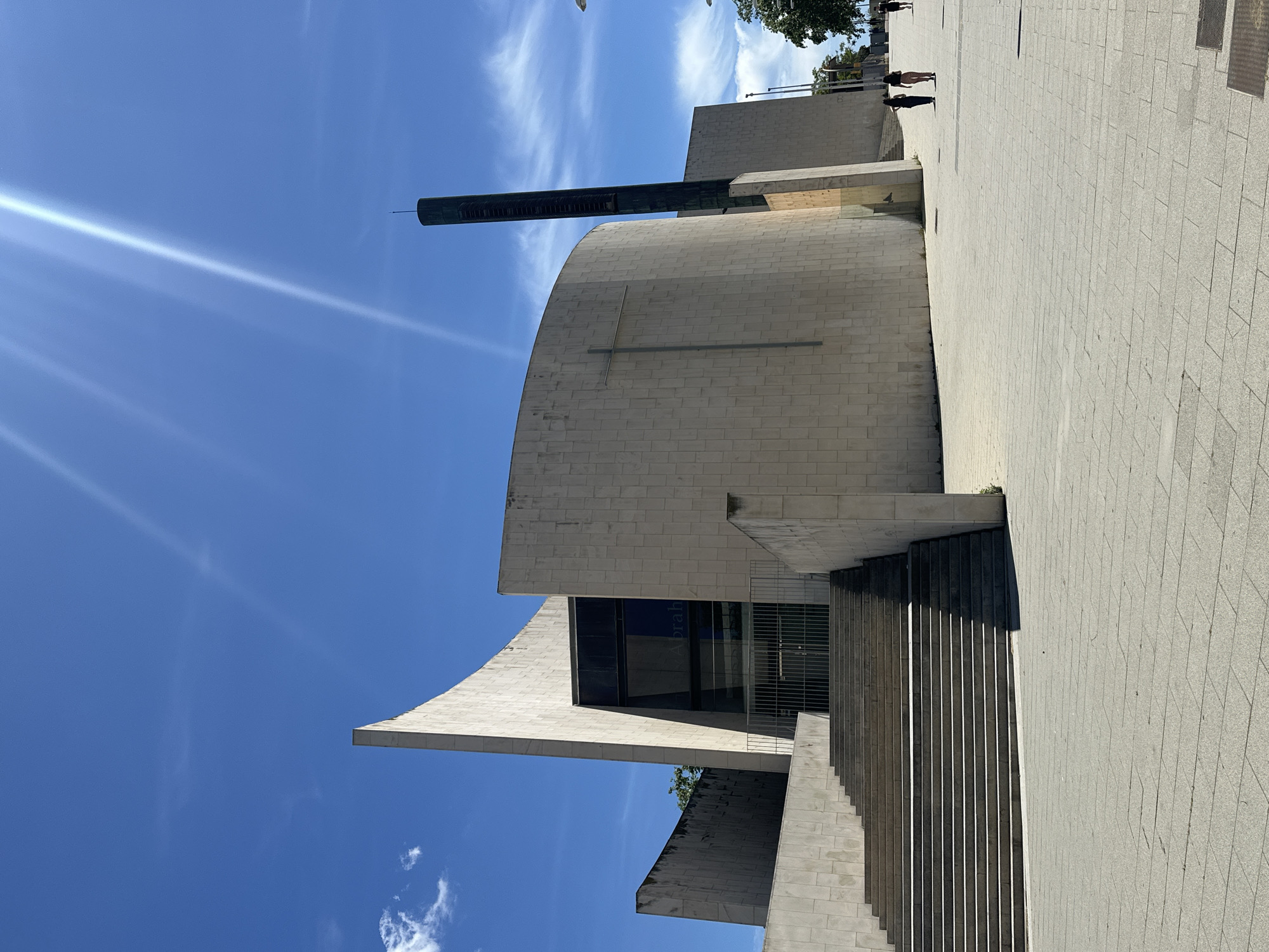 The image shows the Basilica de Sagrat Cor, a church located on the summit of Mount Tibidabo in Barcelona, Spain. The building features a pale gray brick exterior with a curved facade and a tall, slender tower extending from the top. A cross is affixed to the facade. A staircase leads up to an entrance of the building. The surrounding area includes a paved walkway and hints of greenery, suggesting a park-like environment. A few people are visible in the background. The sky is blue with scattered clouds.