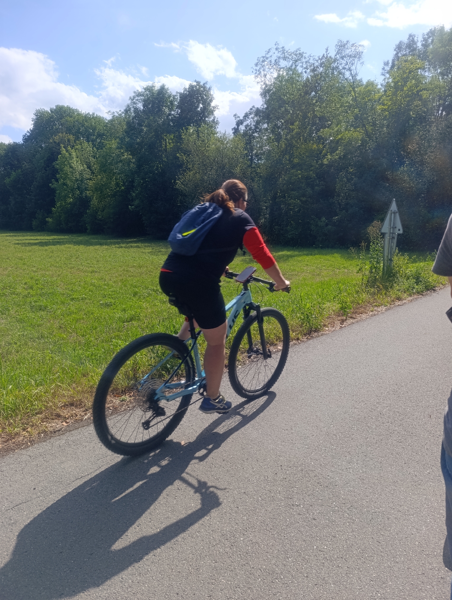 A person is riding a light blue bicycle on a paved path. The rider is wearing a black t-shirt with red sleeves, black shorts, and blue athletic shoes. They have a blue backpack with yellow accents on their back. The bicycle has black tires and a light blue frame. The person is casting a shadow on the path.
The environment is a grassy field with a line of trees in the background. The sky is blue with scattered white clouds. A signpost is visible near the trees. The paved path appears to be a dedicated bike or pedestrian path. The lighting suggests it is daytime, likely late morning or early afternoon, given the length and angle of the shadow.
This scene is likely a park or a recreational area in or around the city of Olomouc, Czechia, given the context provided, as it is a common type of landscape for such areas.