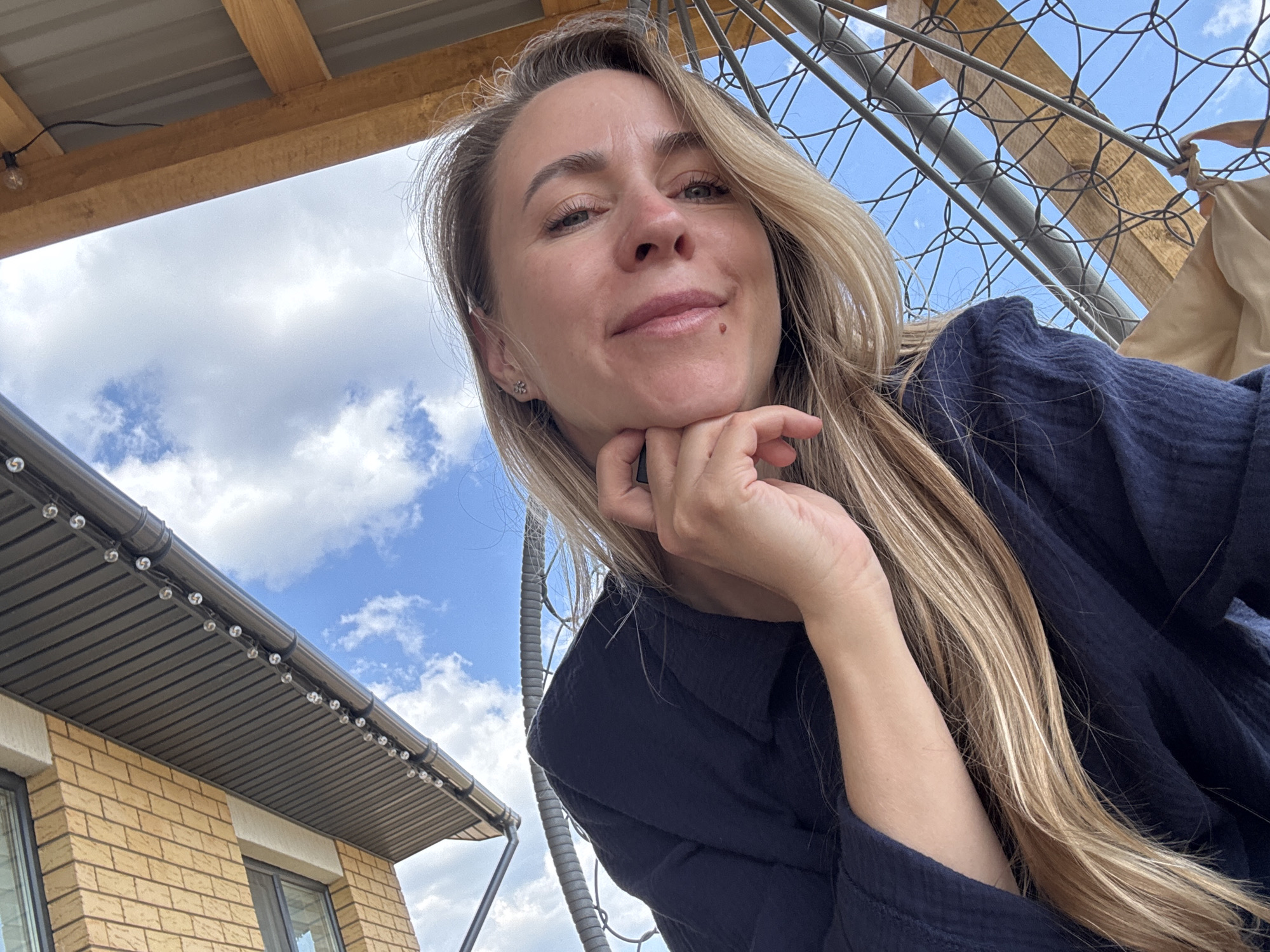A woman with light brown hair poses for a selfie against a backdrop of a partly cloudy sky. She is wearing a dark blue, textured long-sleeved shirt and has her hand resting under her chin. To the upper left is a wooden structure with a string of lights attached. To the lower left is a brick building with dark trim and windows. To the upper right is a chain-link fence. This image was taken in the city of Chaikovsky, Russia.