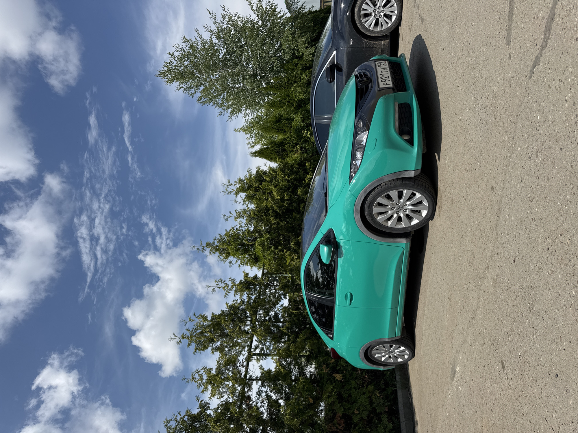 A teal Honda Civic is parked in a lot with a black car behind it. The cars are parked in front of a line of tall evergreen trees and a few deciduous trees. The sky is blue with scattered white clouds. The ground is paved with asphalt. The license plate on the teal Honda Civic is visible and reads "P921TH 159". This image was taken in the city of Chaikovsky, Russia.