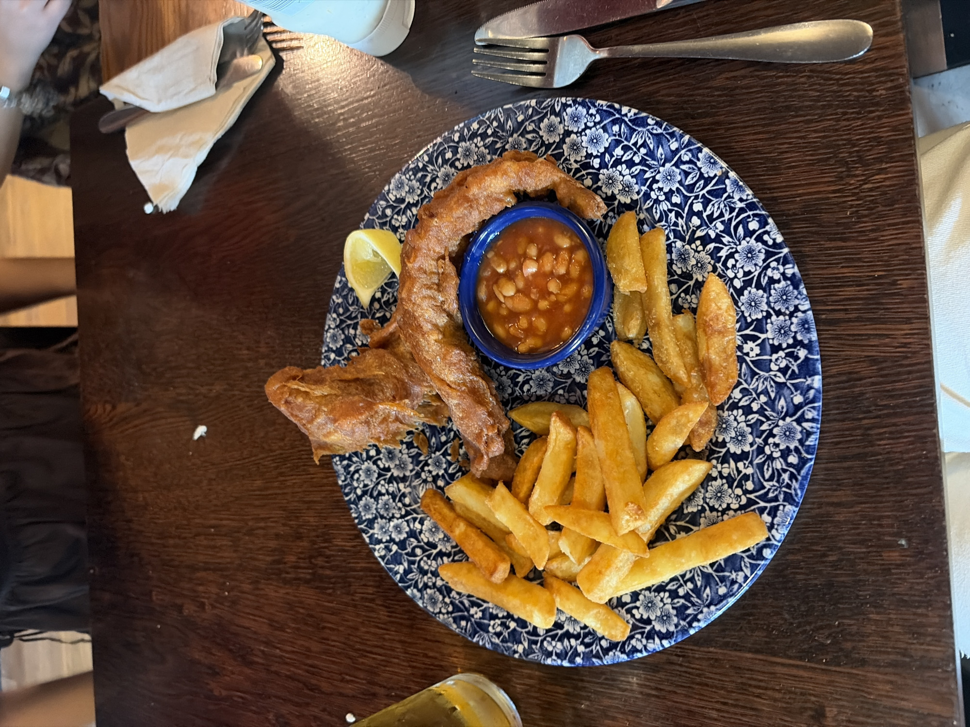 A top-down shot shows a plate of fish and chips on a wooden table. The plate is blue and white with a floral pattern. The fish, which looks like a battered and fried fish, sits on one side of the plate next to a lemon wedge. In the center of the plate is a small blue bowl filled with baked beans. Chips (french fries) surround the fish and beans.
To the left of the plate is a cutlery set wrapped in a napkin and part of a person is visible. A fork and knife are on the right. A glass containing a yellow liquid is visible in the bottom left corner of the image.