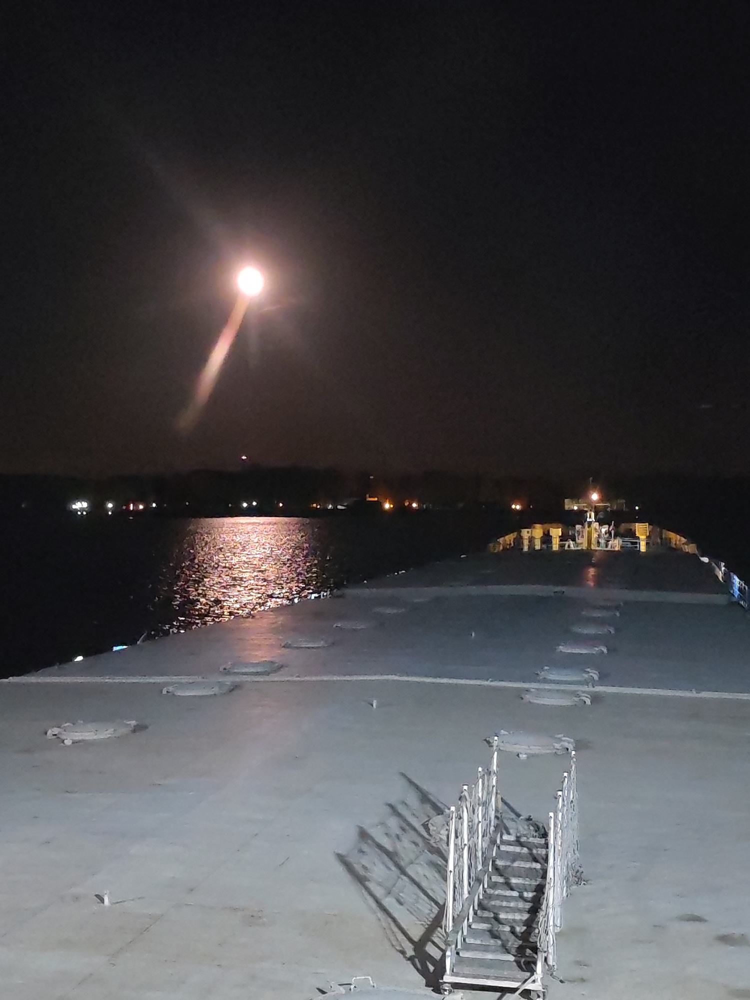The image shows a nighttime scene on what appears to be the deck of a ship or large vessel. The deck is flat and gray, with hatches and other fixtures visible. A set of portable stairs is positioned on the deck, casting long shadows. In the background, there's a body of water reflecting the moonlight. Beyond the water, a dark land mass is visible, dotted with faint lights. Above, the moon shines brightly, casting a strong beam of light. The city and location are undetermined.