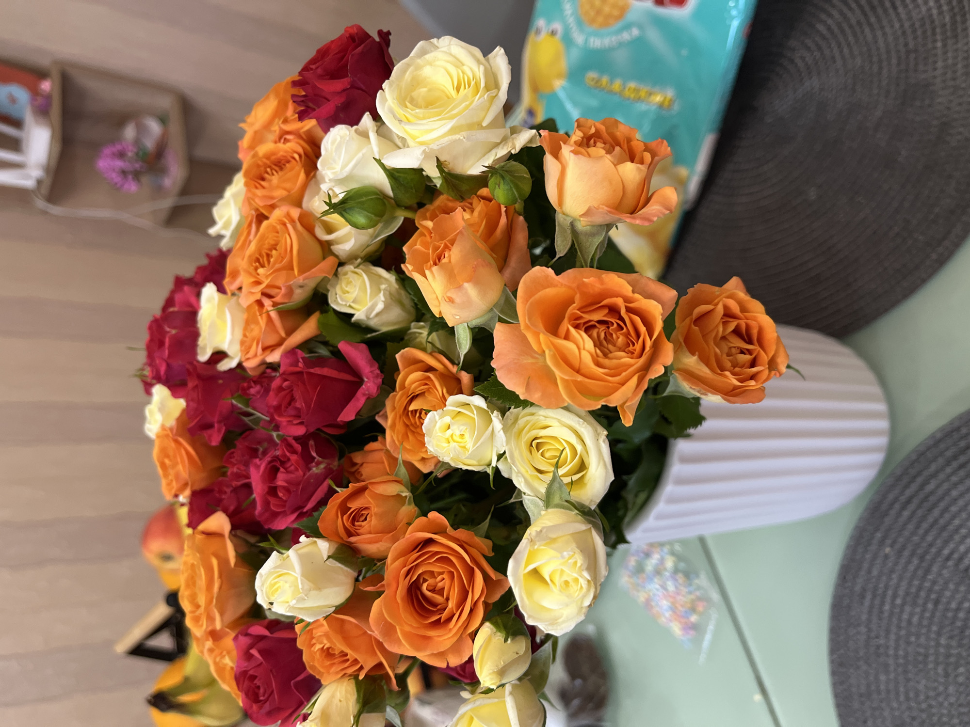 A close-up, high-angle shot shows a white, ribbed vase overflowing with a vibrant bouquet of roses. The roses are a mix of colors, including deep red, bright orange, and soft cream. Some buds are visible, along with green leaves. The vase sits on a light green surface, possibly a table, with a textured dark gray placemat in the foreground. In the background, there are hints of a kitchen or dining area with shelves holding decorative items, including a small purple floral arrangement. A bag of brightly colored snacks is visible to the right of the vase. The overall impression is one of a cheerful and inviting domestic setting. There are no people or animals visible in the image. The image does not provide any information about the city of Sochi, Russia.