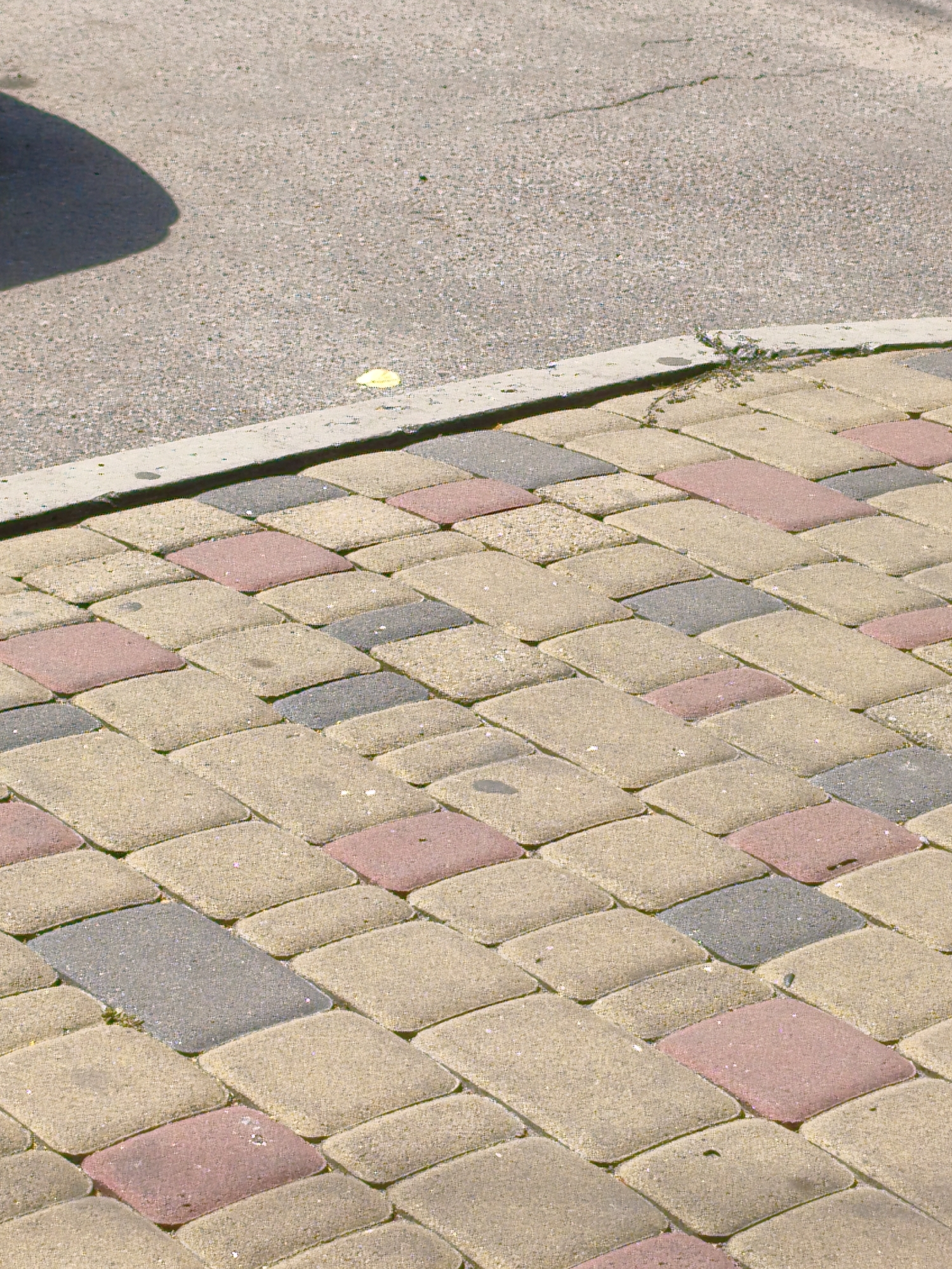 The image shows a close-up, angled view of a paved sidewalk made of interlocking pavers in a herringbone pattern. The pavers are of three colors: beige, pinkish-red, and gray. The sidewalk is bordered by a concrete curb. Behind the curb, a section of asphalt pavement is visible, with a shadow of an unseen object casting across it from the left. The asphalt appears to have a crack running through it. A small yellow leaf or piece of debris is visible on the asphalt near the curb. The scene is outdoors and appears to be a street or walkway. The location is identified as Velyka Bahachka, Ukraine.