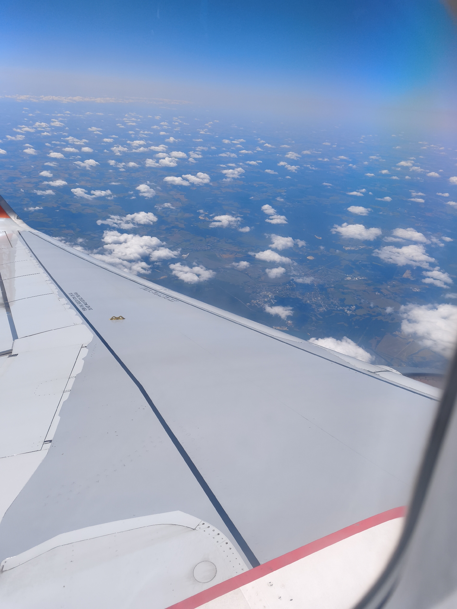 An aerial view from an airplane window shows a landscape below with scattered clouds and a portion of the airplane wing in the foreground. The wing is white with a red stripe along its edge. Below, the land is a patchwork of green fields and darker forested areas, with a town visible in the distance. The sky is a clear, bright blue. The town, identified as Trebbin, Germany, is spread out with numerous buildings, roads, and appears to be bordered by a river or body of water.