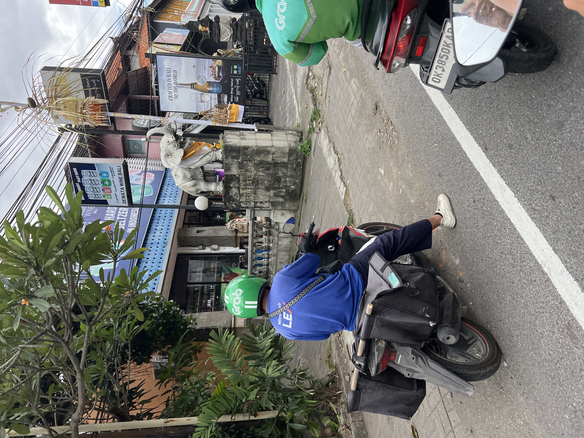 In Denpasar, Indonesia, a male Grab delivery driver wearing a green helmet and a blue long-sleeved shirt with "LEX" printed on the back rides a red and black motorcycle with large black saddlebags. He is stopped on a street next to a sidewalk with a white and orange striped demarcation line. Behind him, another Grab delivery driver on a red motorcycle is partially visible. In the background, there is a building with a blue facade and windows, a grey elephant statue on a pedestal, and various signs including one advertising "CRAFTED IN BALI TO THE WORLD". Overhead power lines are strung across the sky. The weather appears overcast.