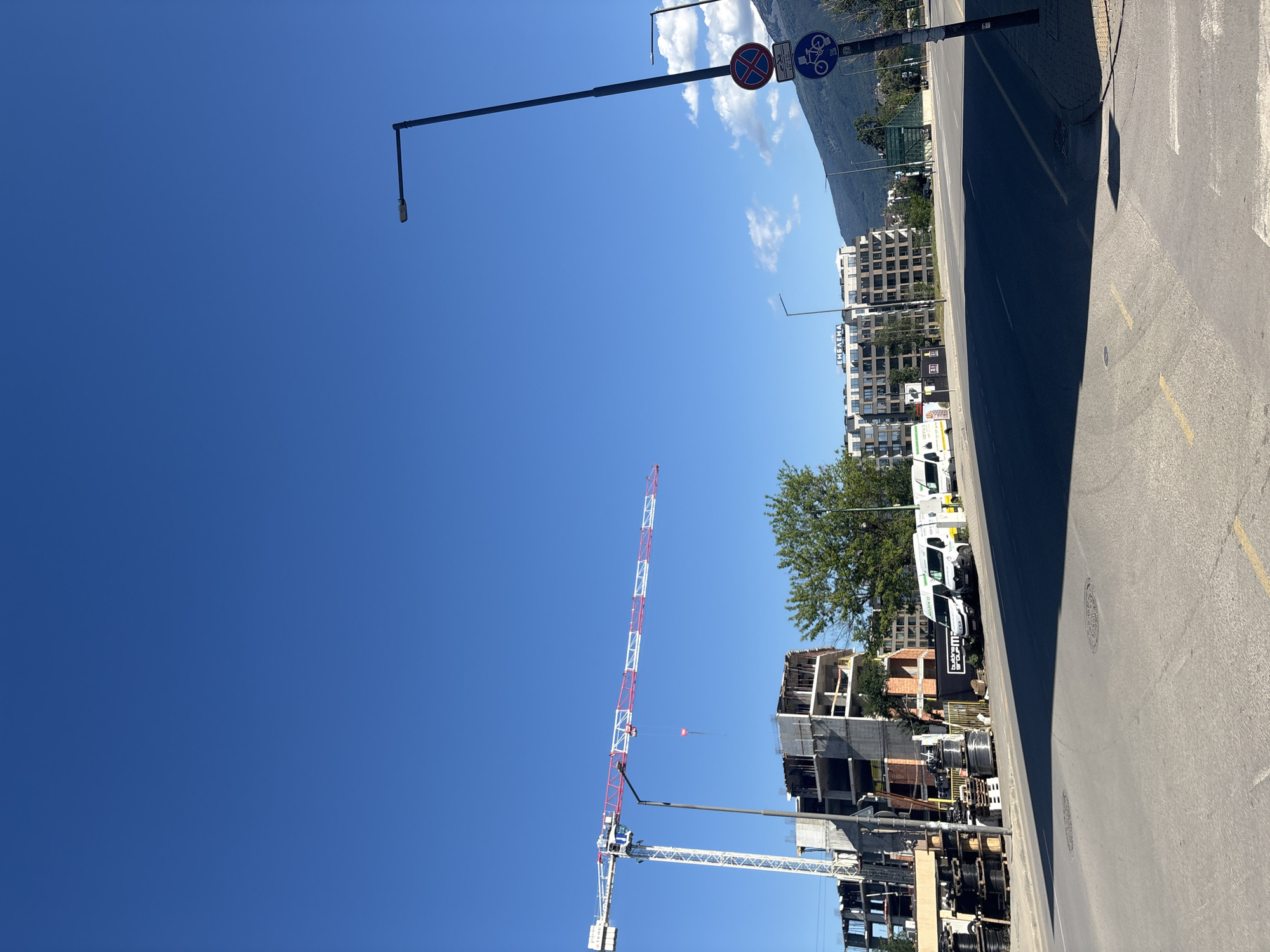 A construction site is visible on the left side of the frame, featuring a tall white and red crane with a partially constructed building beneath it. The building appears to be in its early stages of development, with exposed rebar and concrete. Several vans are parked in front of the construction site. In the background, a modern apartment building with a sign reading "EMELENA" is visible, along with a forested mountain range. A street runs through the foreground, casting a shadow across the asphalt. Traffic signs, including a no parking sign and a bicycle lane sign, are present on the right side of the street. The sky is clear and blue.