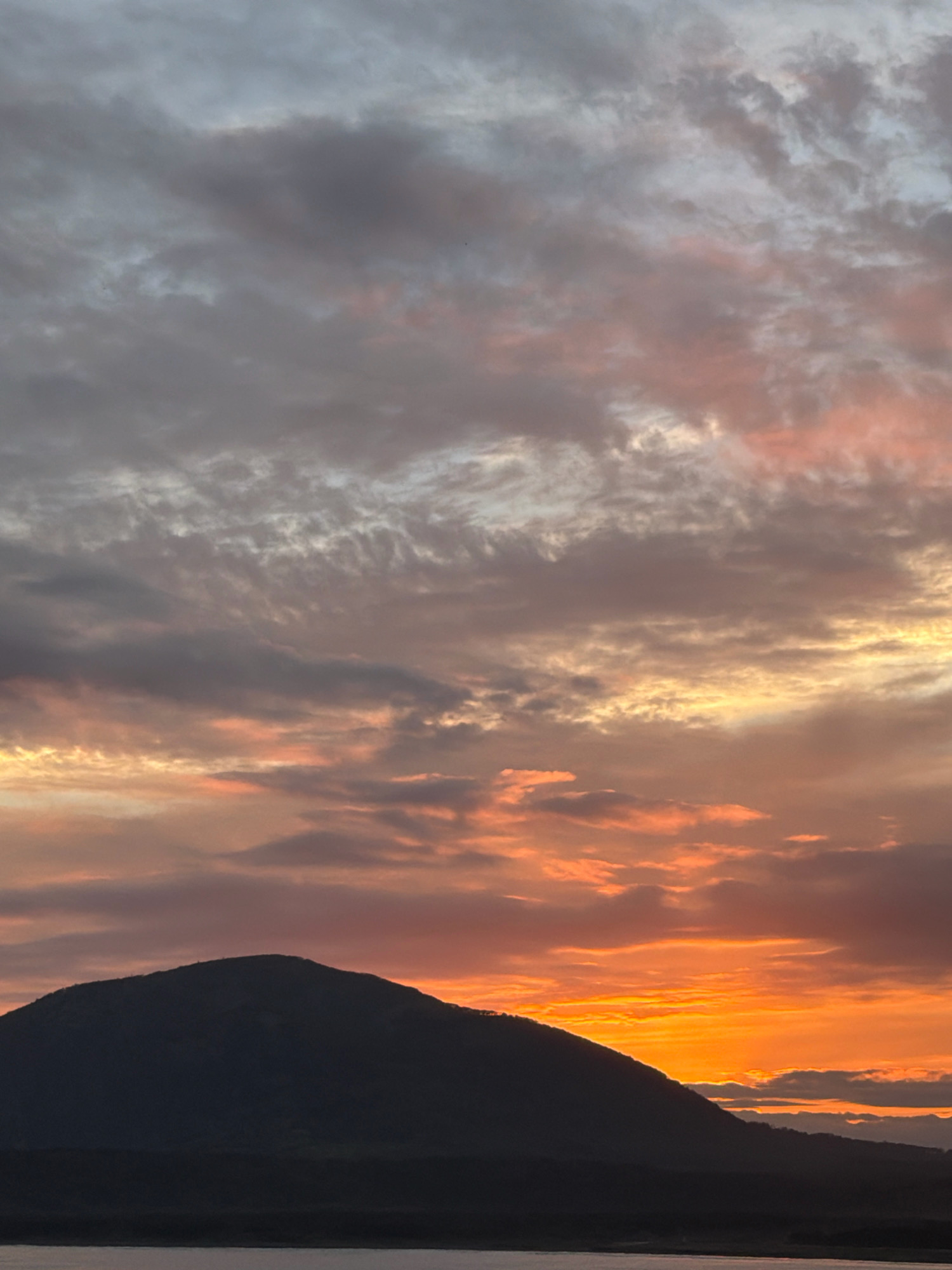 A sunset scene over a body of water in Yuzhno-Kurilsk, Russia. The sky is filled with dramatic clouds, ranging from soft grays and pinks near the top to vibrant oranges and yellows closer to the horizon. A dark, silhouetted mountain range dominates the lower portion of the image, with its peak obscuring the setting sun. The water in the foreground is calm and reflects the colors of the sky.
