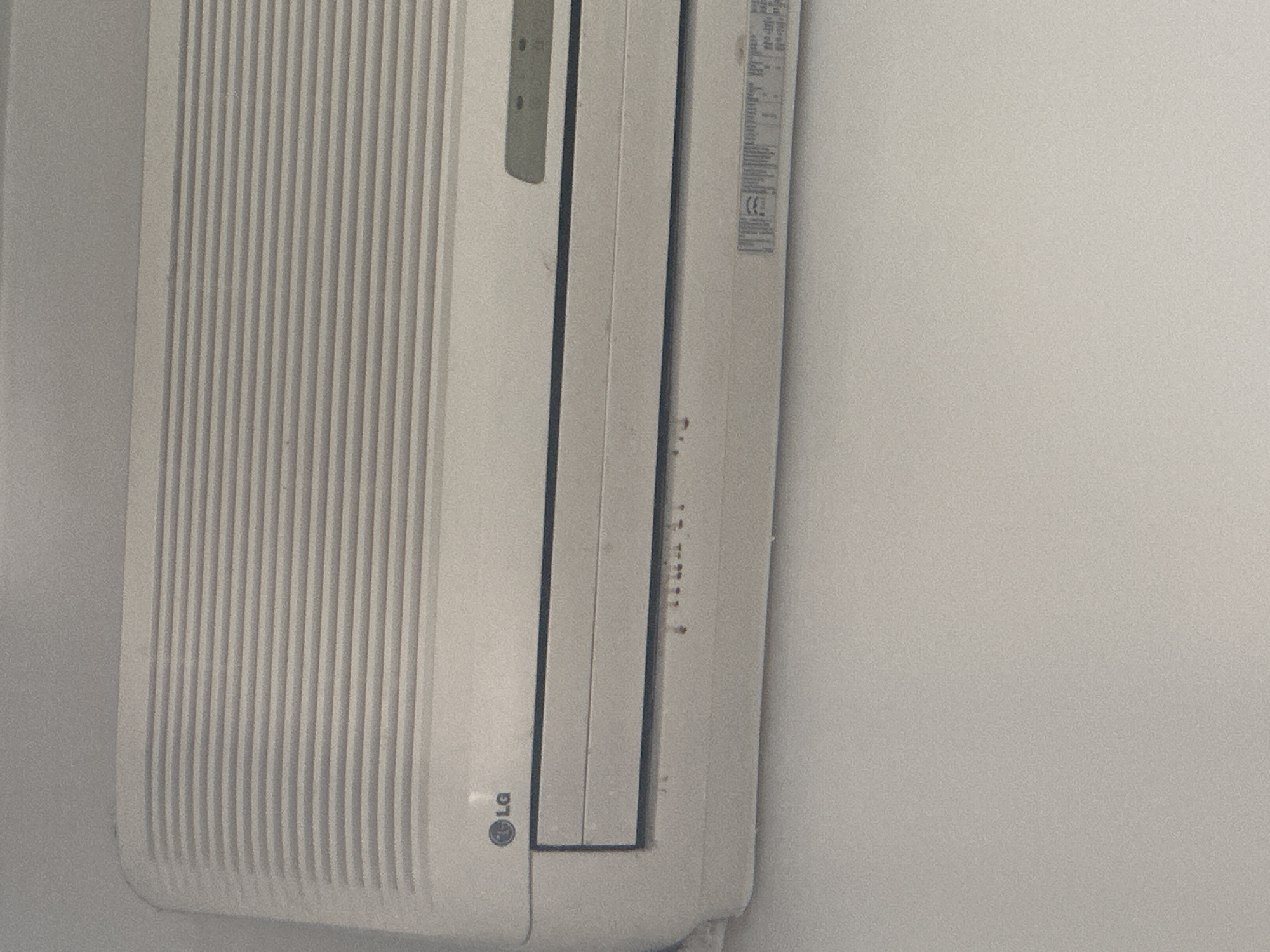 This is a close-up, vertical, eye-level shot of a white LG air conditioner unit mounted on a wall. The unit has a textured surface with vertical grooves on the left side. A portion of a silver-colored label with some text and a circular CE mark is visible on the right side of the unit, partially obscured by dirt. The overall appearance of the unit suggests it may be an indoor air conditioner. The lighting is diffused and even, with no strong shadows, indicating an indoor setting with ambient light. The wall behind the unit is a plain, light-colored surface, likely painted. The image is slightly grainy, suggesting it was taken with a smartphone or a camera in low light conditions. There is some visible dirt and smudging on the unit's surface, particularly on the right side where the label is located. The scene is static, with no people or activity depicted. The environment appears to be a residential or commercial indoor space.